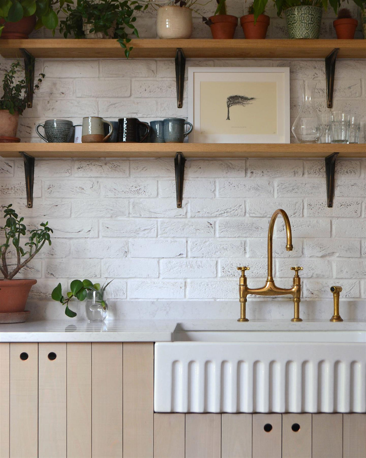 A straight-on view of a contemporary kitchen with rustic beech cabinetry, a fluted Belfast sink, white metro wall tiles and oak shelving with classic utilitarian shelf brackets in a dark oxidised brass finish. TEST