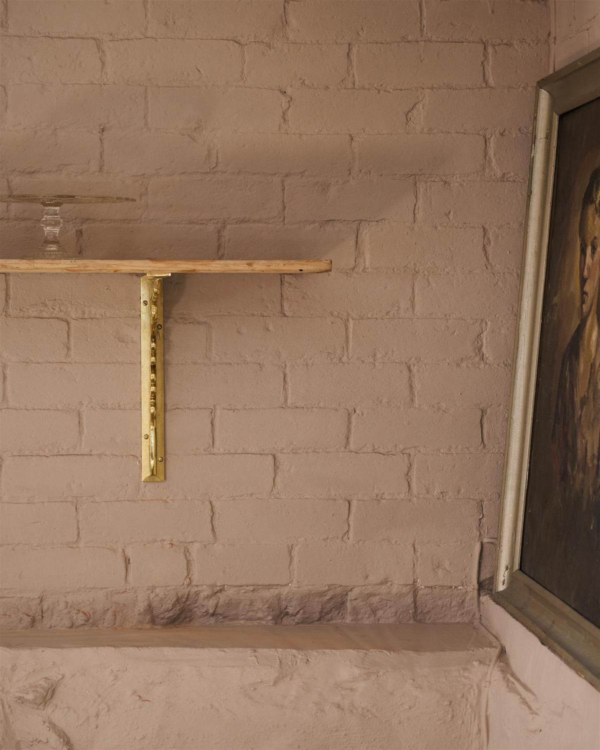 A straight-on view of a large Victorian-inspired ornate aged brass shelf bracket holding a deep pine shelf to a pink colour brick wall. TEST