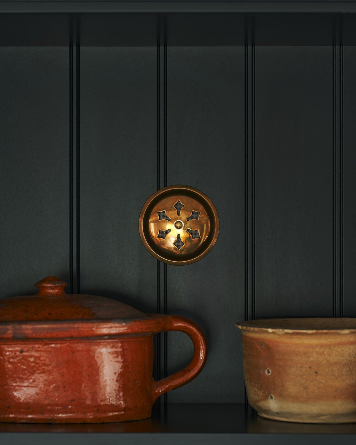 A Victorian-inspired circular brass ventilation grille with traditional decorative cutouts fitted to a black painted tongue and groove-style cupboard back panel. TEST