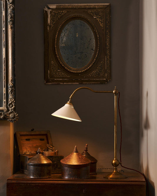 An oblique view of an antique table with a Victorian-inspired desk lamp with a solid brass foot, stand, curved arm and a small cream glazed ceramic light shade. A red twisted fabric flex cord can be seen coming from the top finial.