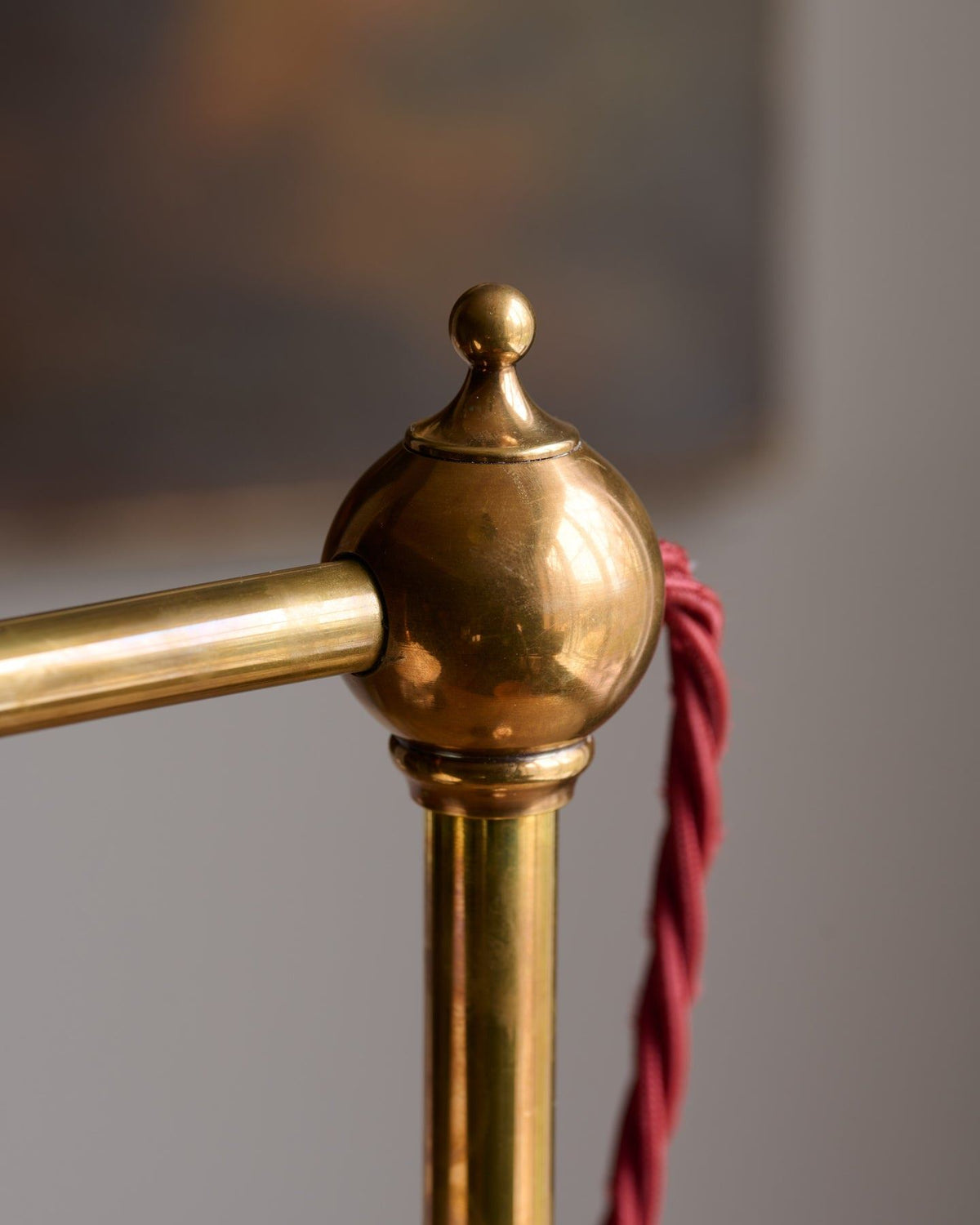 An oblique view of a Victorian-inspired desk lamp with a solid brass foot, stand, curved arm and a small cream glazed ceramic light shade. A red twisted fabric flex cord can be seen coming from the top finial. TEST