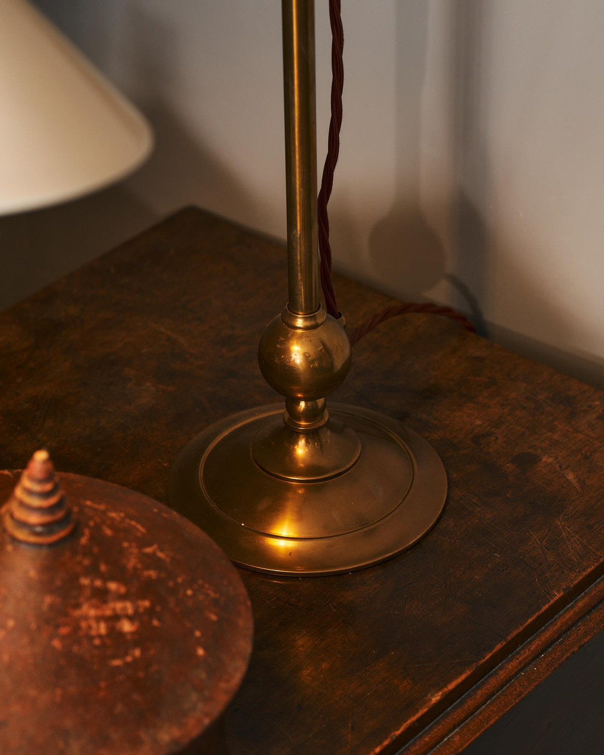 A straight-on view of an antique table in front of a pink painted glazed partition wall. On the table is a Victorian-inspired desk lamp with a solid brass foot, stand, curved arm and a small cream glazed ceramic light shade. TEST