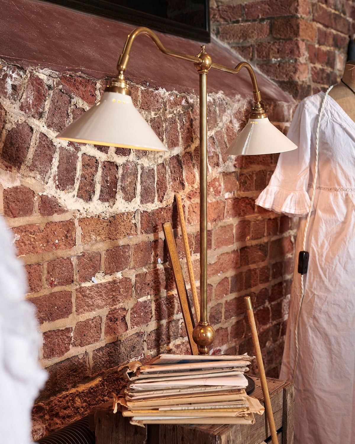 An oblique view of a Victorian-inspired desk lamp with a solid brass foot, stand, two curved arms and two small cream glazed ceramic light shades on a wooden box in front of a red brick wall. TEST