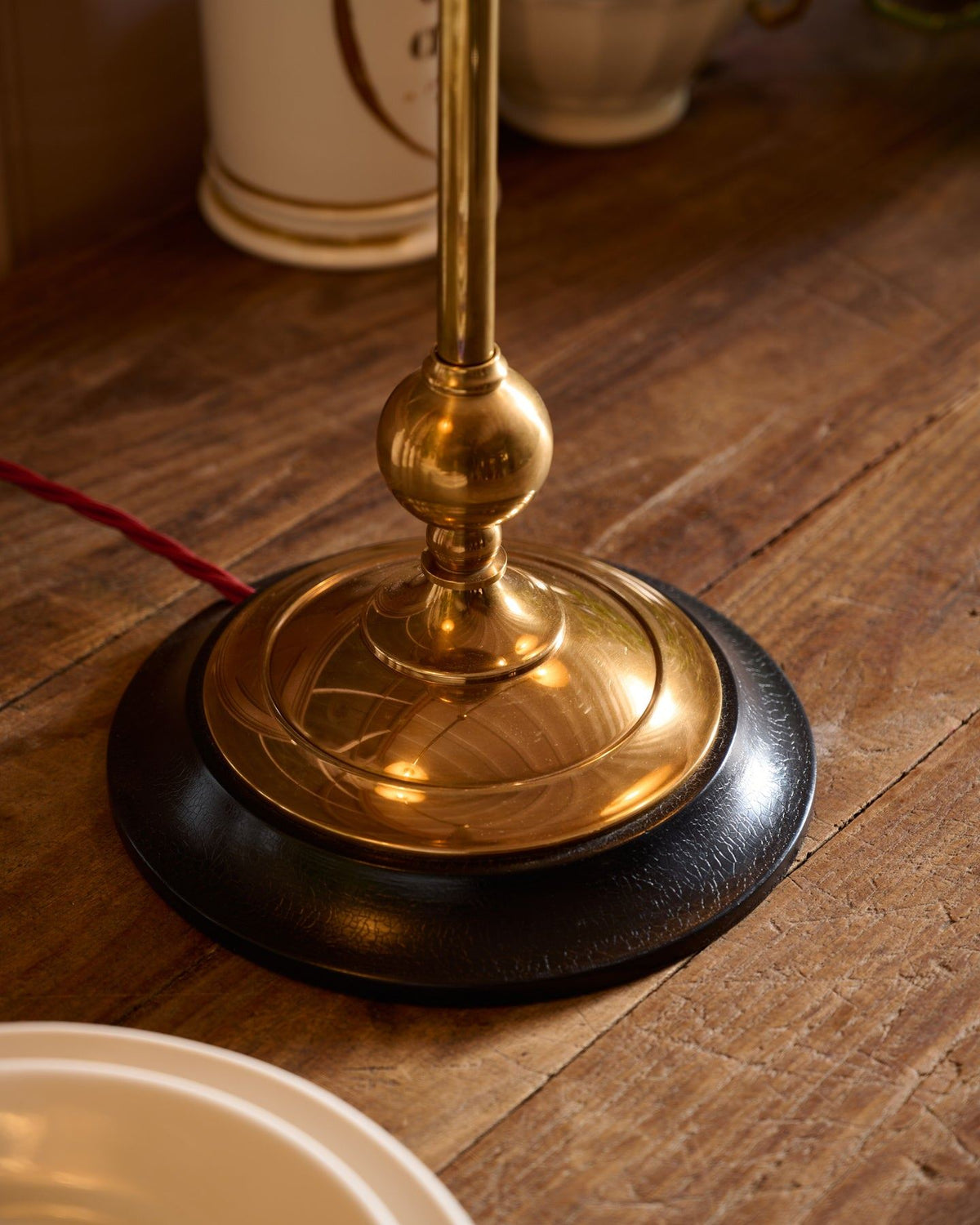 A close-up view of a solid brass desk lamp foot with a dark hardwood base and a red twisted fabric flex cord leading out from one side. TEST