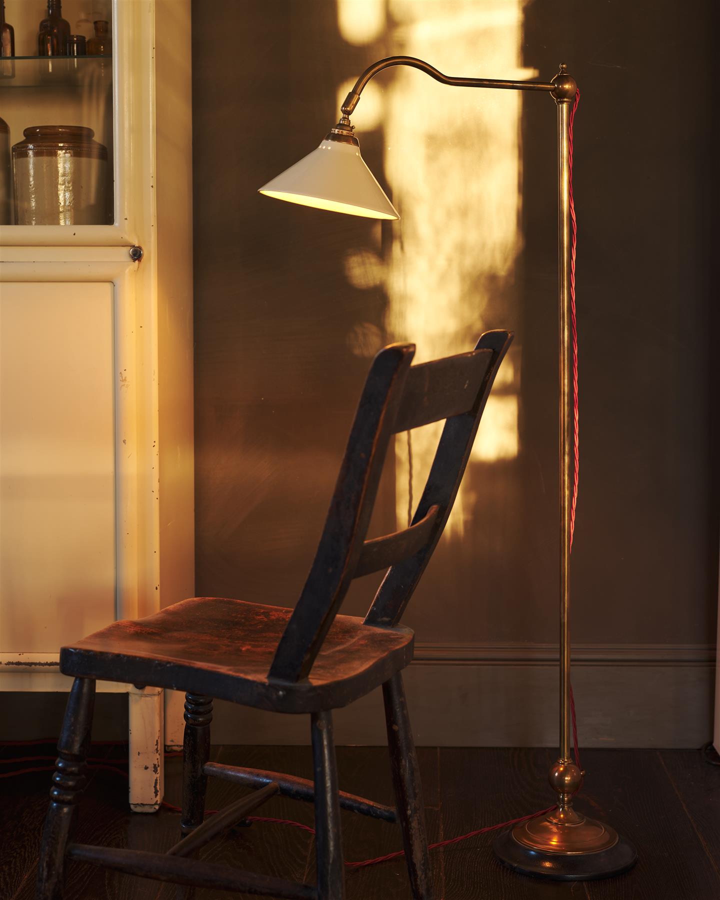 A straight-on view of a Victorian-inspired floor lamp with a solid brass foot, stand, curved arm, dark hardwood base and a small cream glazed ceramic light shade, beside an antique wooden chair, in front of a pink painted glazed partition wall.