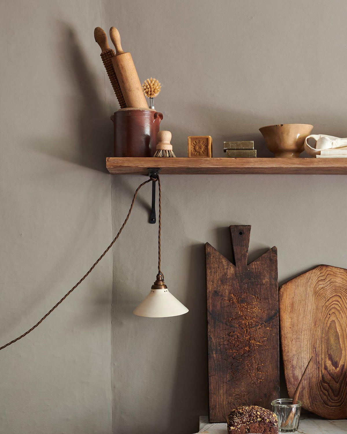 A straight-on view of a small plug-in light with a cream glazed shade, brown twisted fabric flex cord and brass fittings, hanging from an iron shelf bracket under a rustic pine shelf. TEST