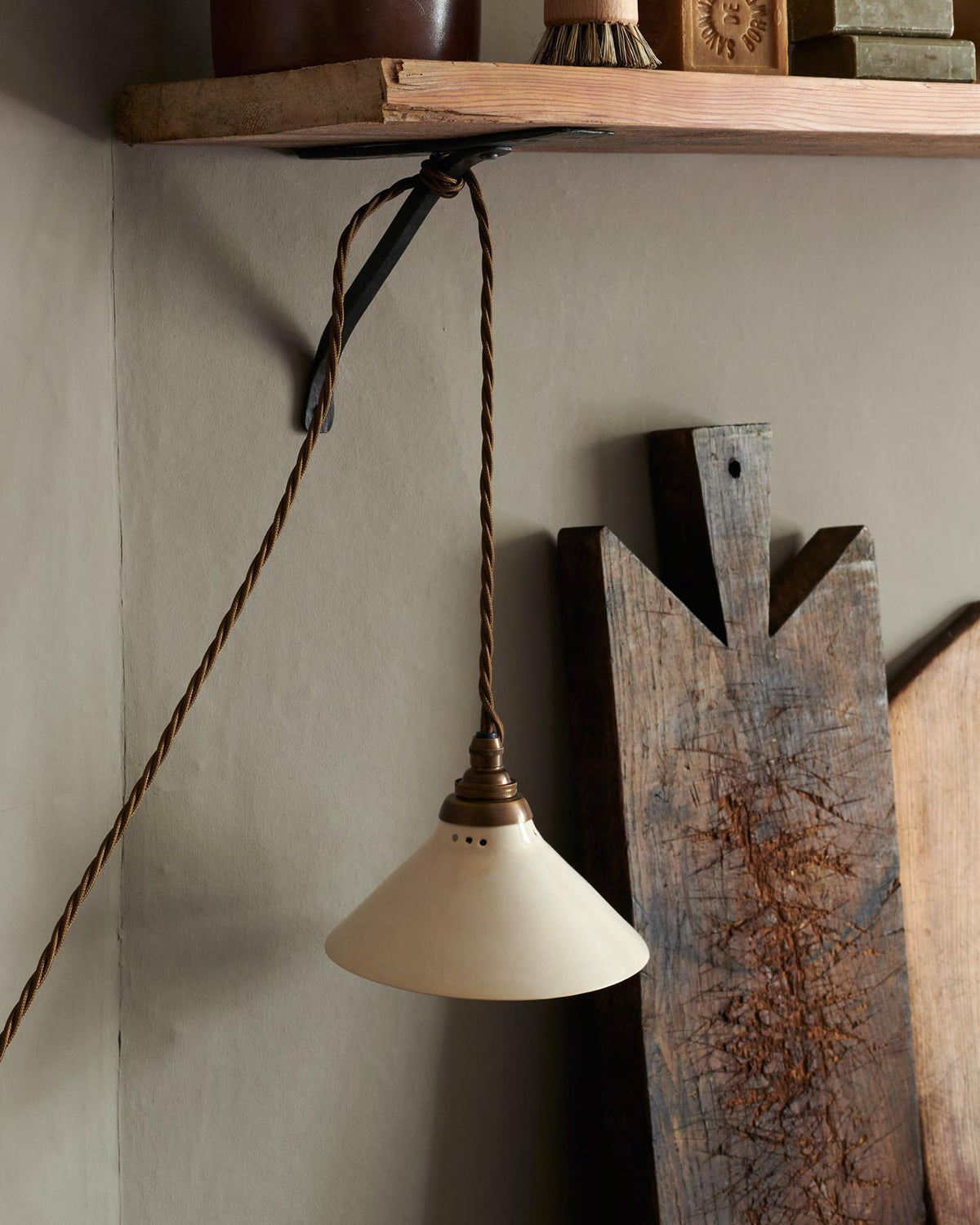 A close-up oblique view of a small plug-in light with a cream glazed shade, brown twisted fabric flex cord and brass fittings, hanging from an iron shelf bracket under a rustic pine shelf. TEST