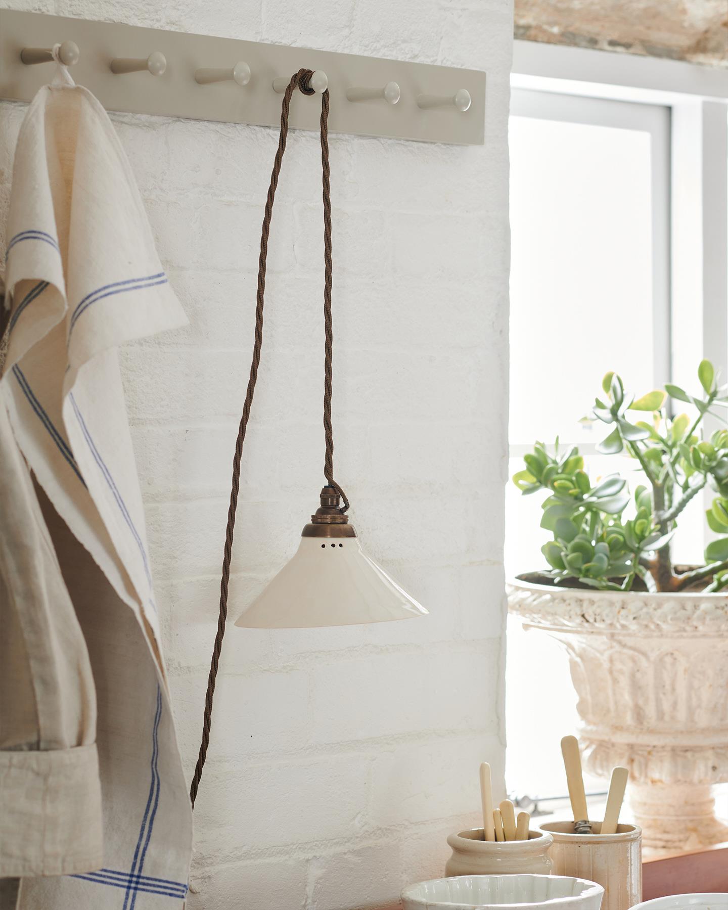 A close-up oblique view of a small plug-in light with a cream glazed shade, brown twisted fabric flex cord and brass fittings, hanging from painted pegs above a kitchen worktop.
