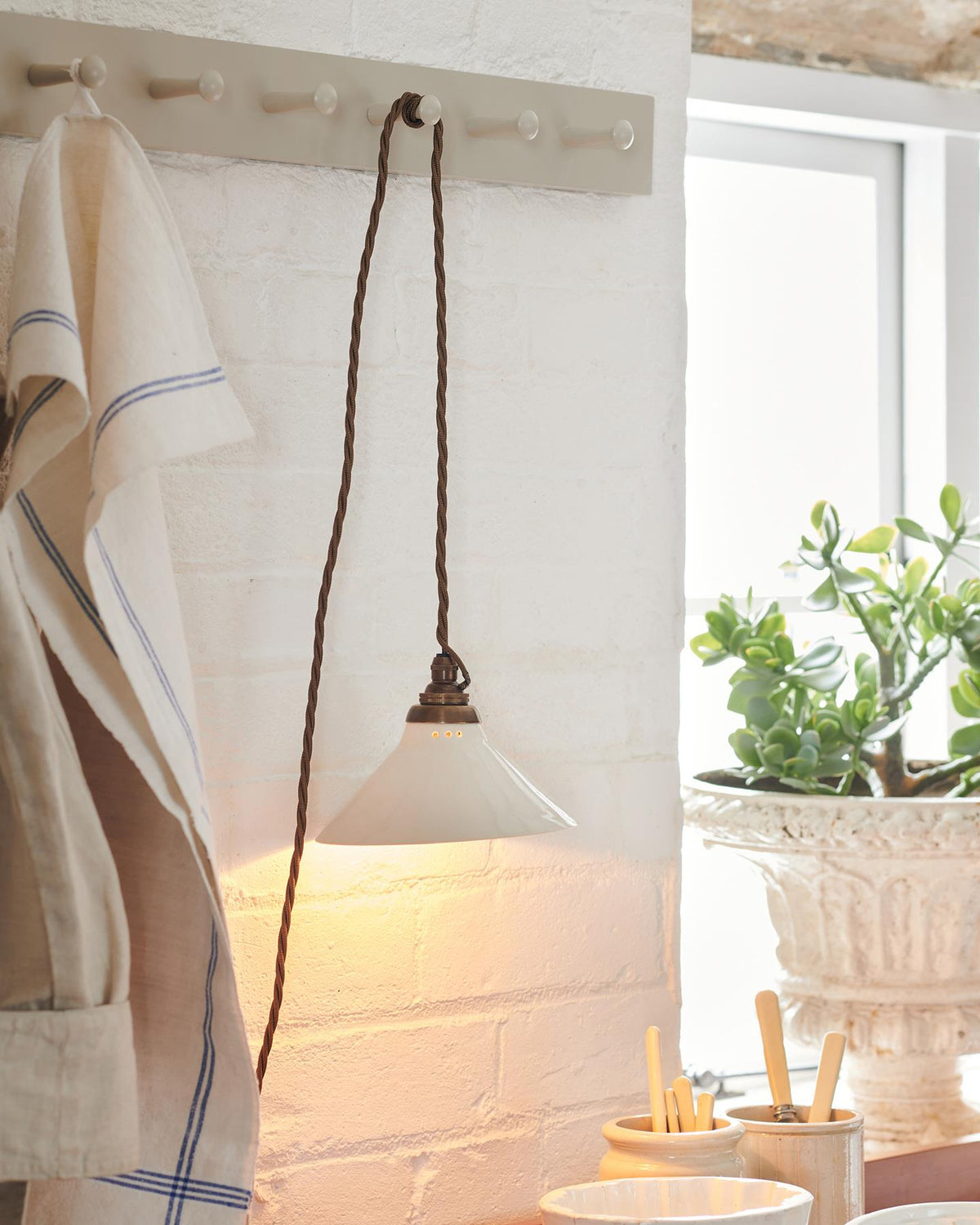 A close-up oblique view of a small plug-in light with a cream glazed shade, brown twisted fabric flex cord and brass fittings, hanging from painted pegs above a kitchen worktop. The light is on. TEST