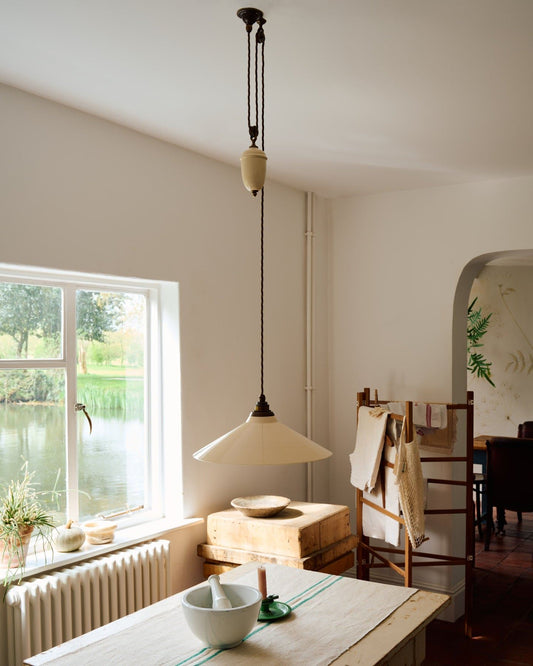 An oblique view of a rise and fall light with a large cream glazed ceramic shade, aged brass fittings and brown twisted fabric flex cord hanging in a white room above an antique pine table.