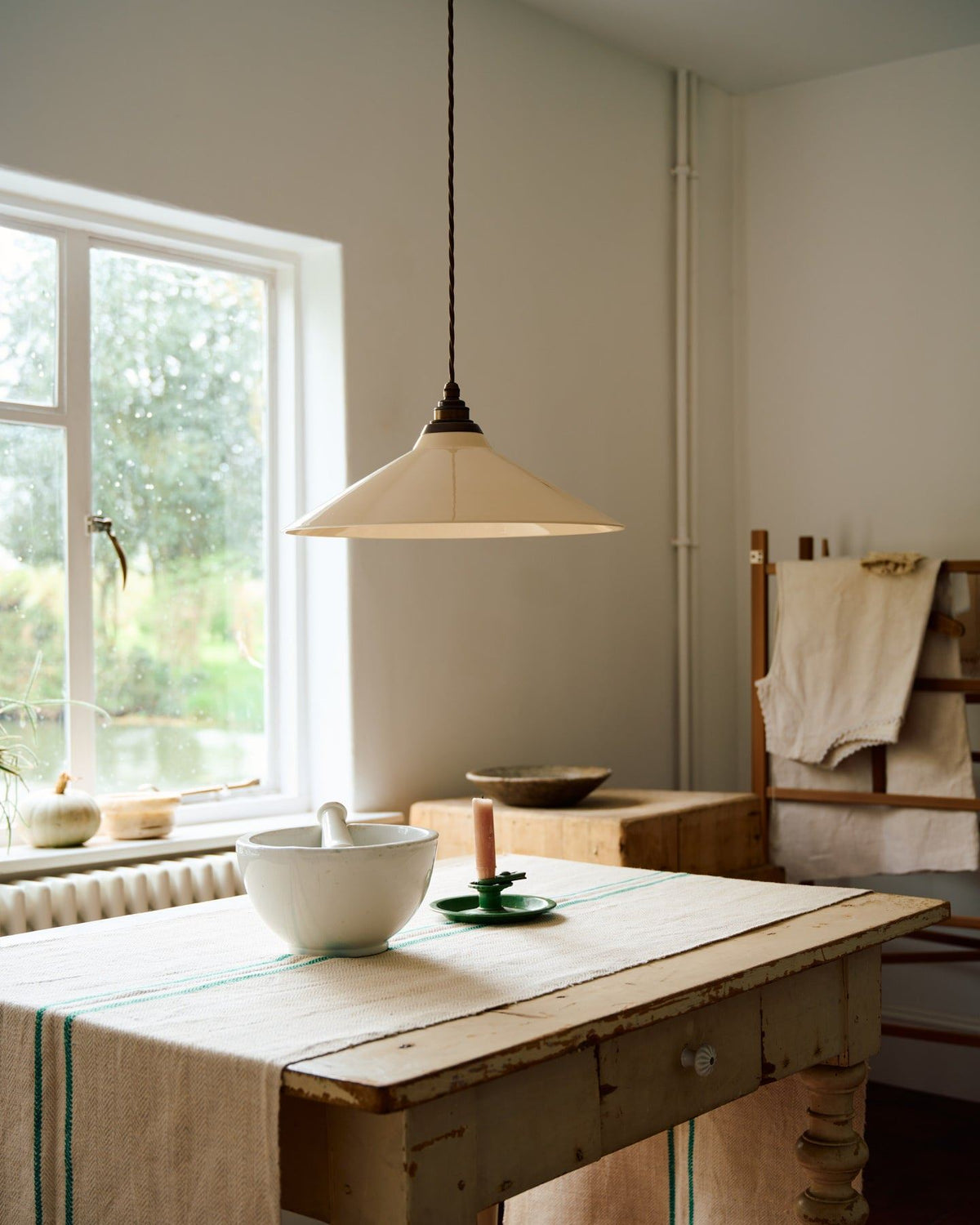 A close-up oblique view of a rise and fall light with a large cream glazed ceramic shade, aged brass fittings and brown twisted fabric flex cord hanging in a white room above an antique pine table. TEST