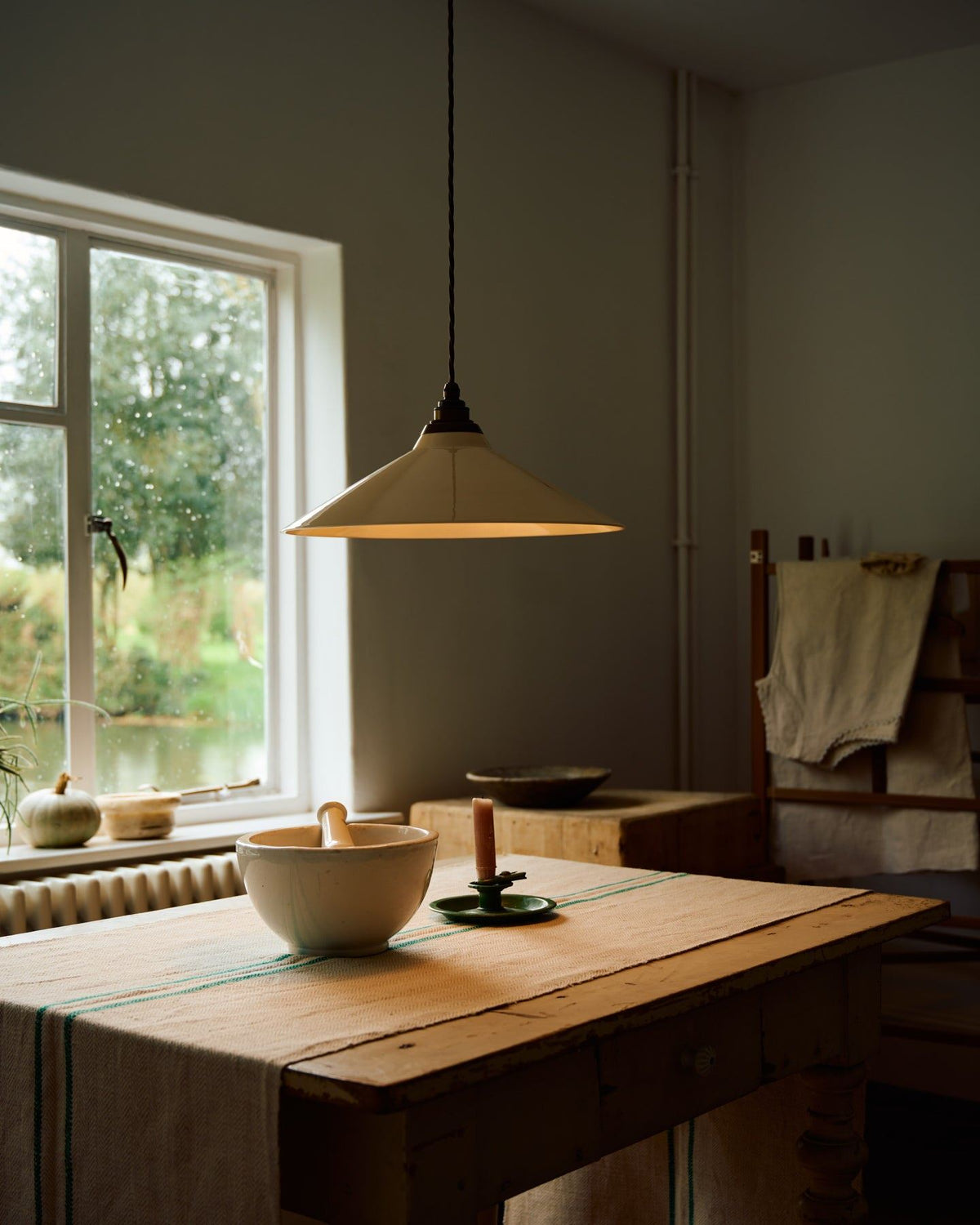 A close-up oblique view of a rise and fall light, turned on, with a large cream glazed ceramic shade, aged brass fittings and brown twisted fabric flex cord hanging in a white room above an antique pine table. TEST