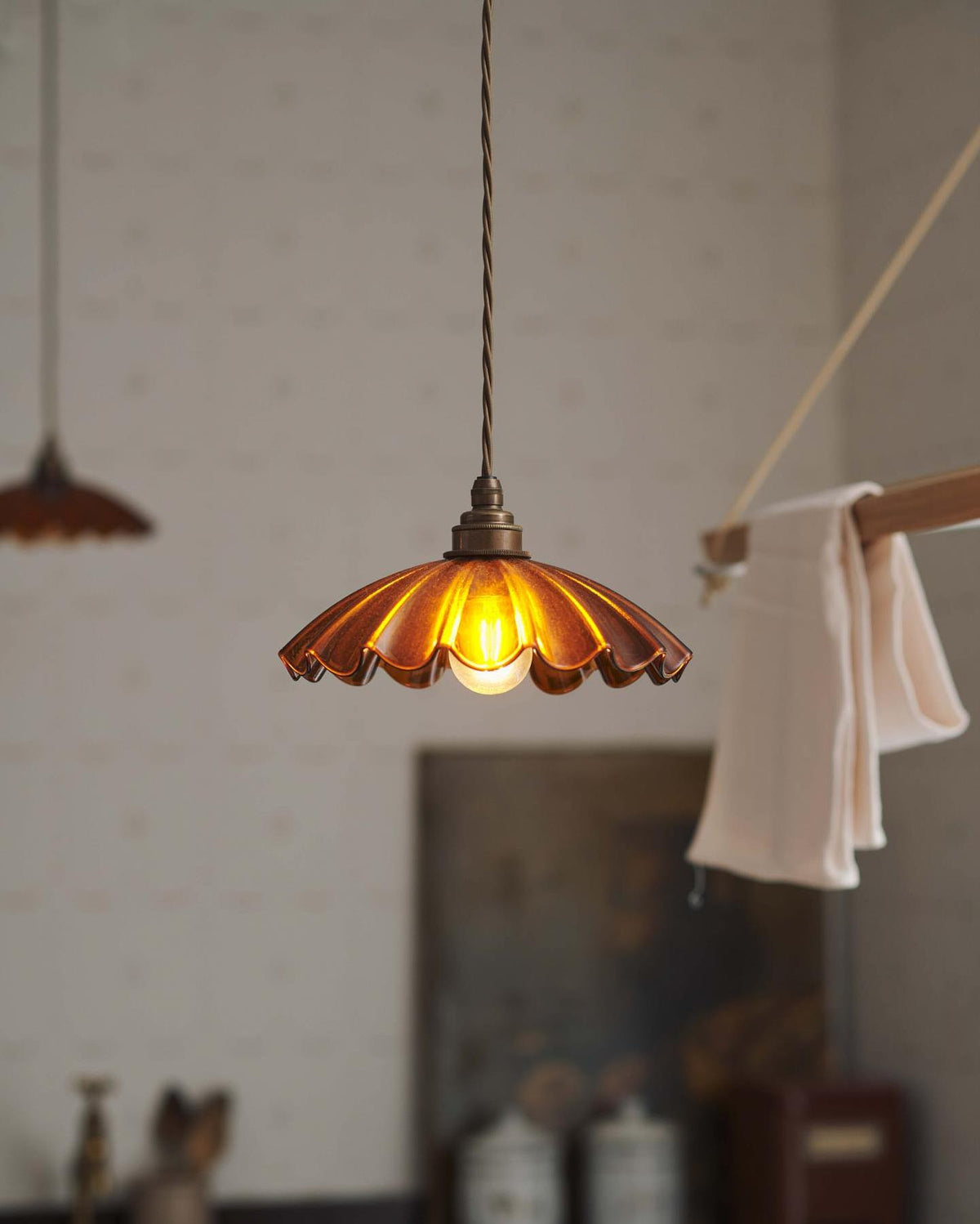 A close-up view of a small frilly amber glass pendant light with brass fittings and brown twisted fabric flex cord. The light is on and shining through the tranlucent glass. TEST