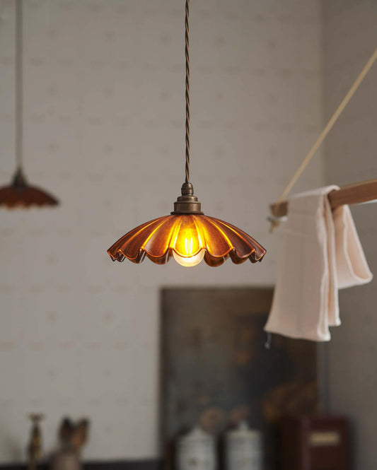 A close-up view of a small frilly amber glass pendant light with brass fittings and brown twisted fabric flex cord. The light is on and shining through the tranlucent glass.