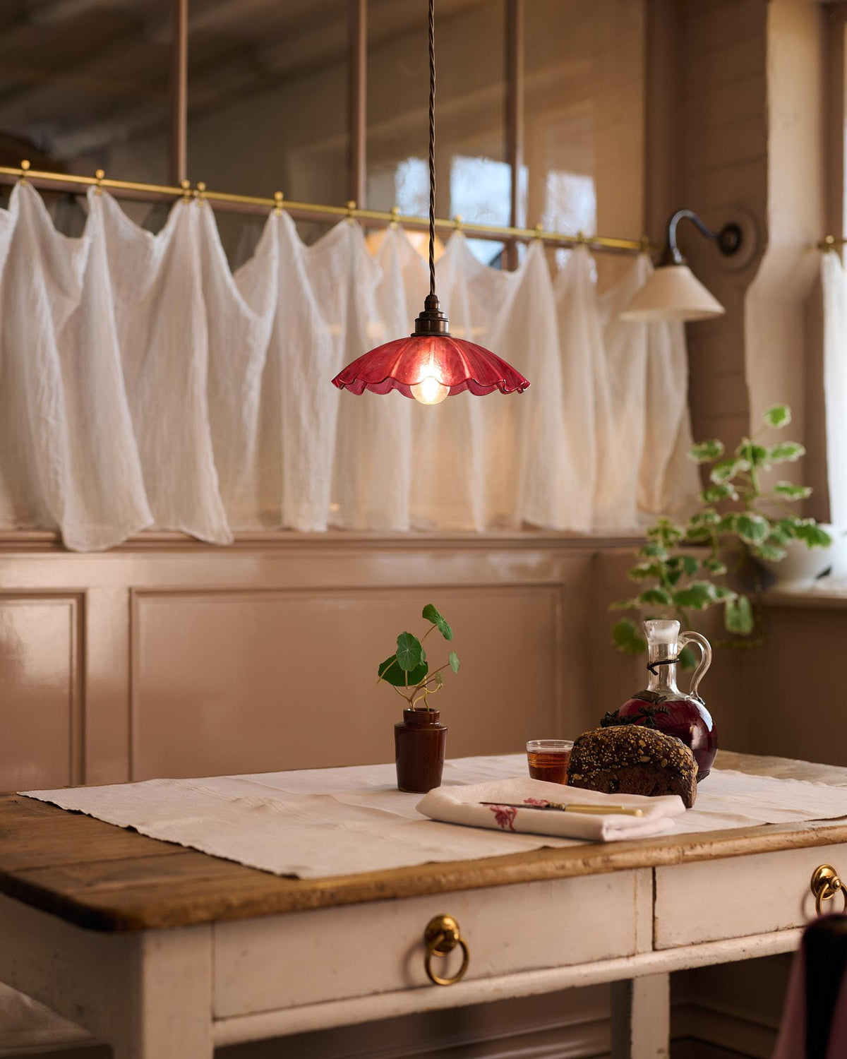 An oblique view of a small frilly cranberry glass pendant light with brass fittings and brown twisted fabric flex cord hanging above an antique pine table in front of a glazed partition with a lace curtain. TEST