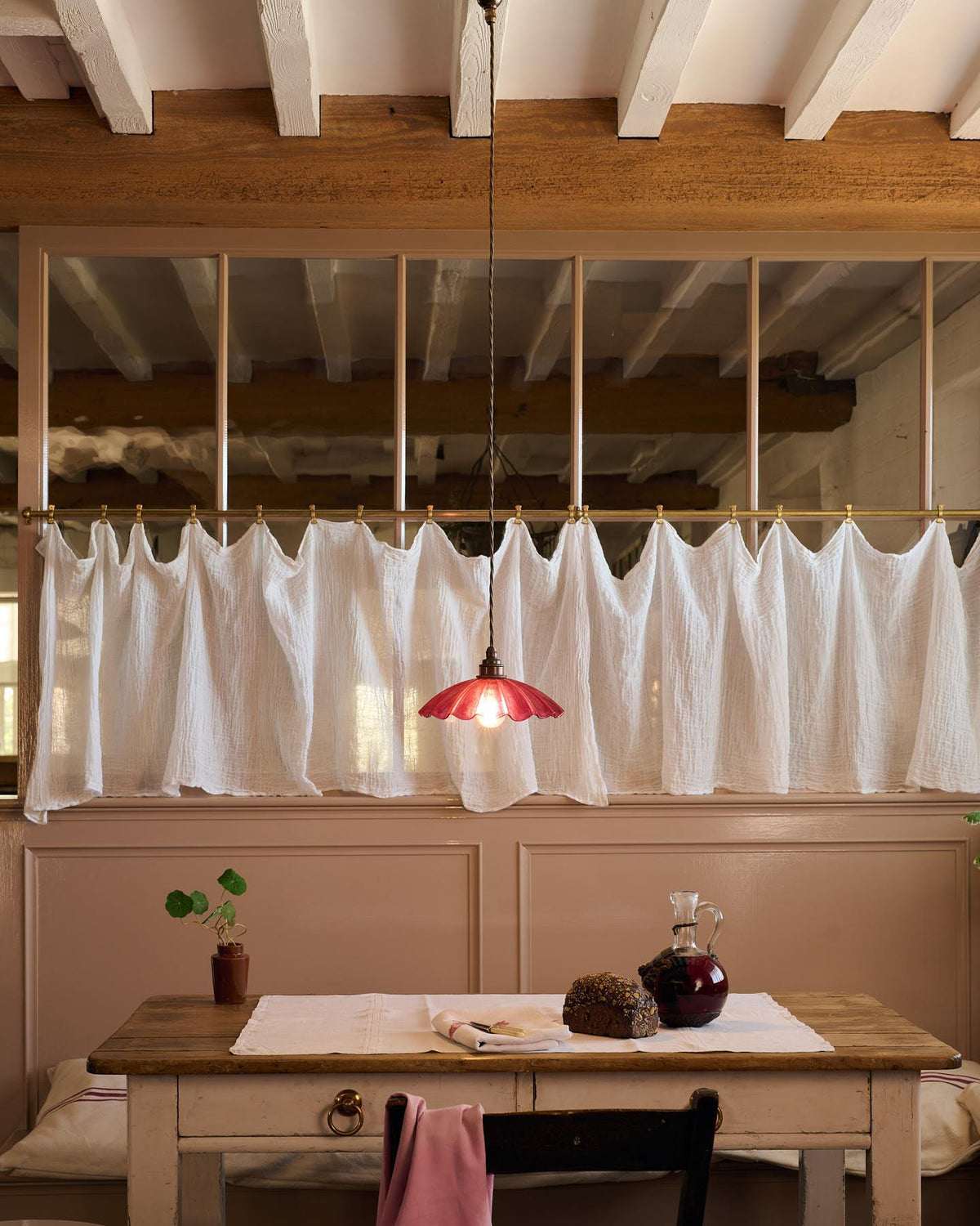 A straight-on view of a small frilly cranberry glass pendant light with brass fittings and brown twisted fabric flex cord hanging above an antique pine table in front of a glazed partition with a lace curtain. TEST