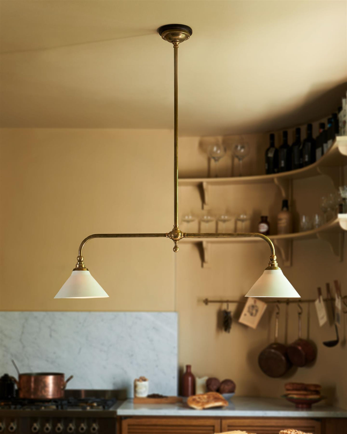 A straight-on view of a mid-century-style kitchen with oak cabinetry, buttery yellow walls and a Victorian-inspired chandelier light with brass arms and small unglazed porcelain shades over a prep table with a Carrara marble worktop. TEST