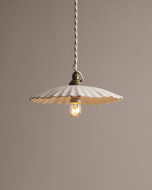 A large narrow pleat porcelain pendant light with an unglazed finish, an aged brass B22 lampholder and a white twisted fabric flex cord.