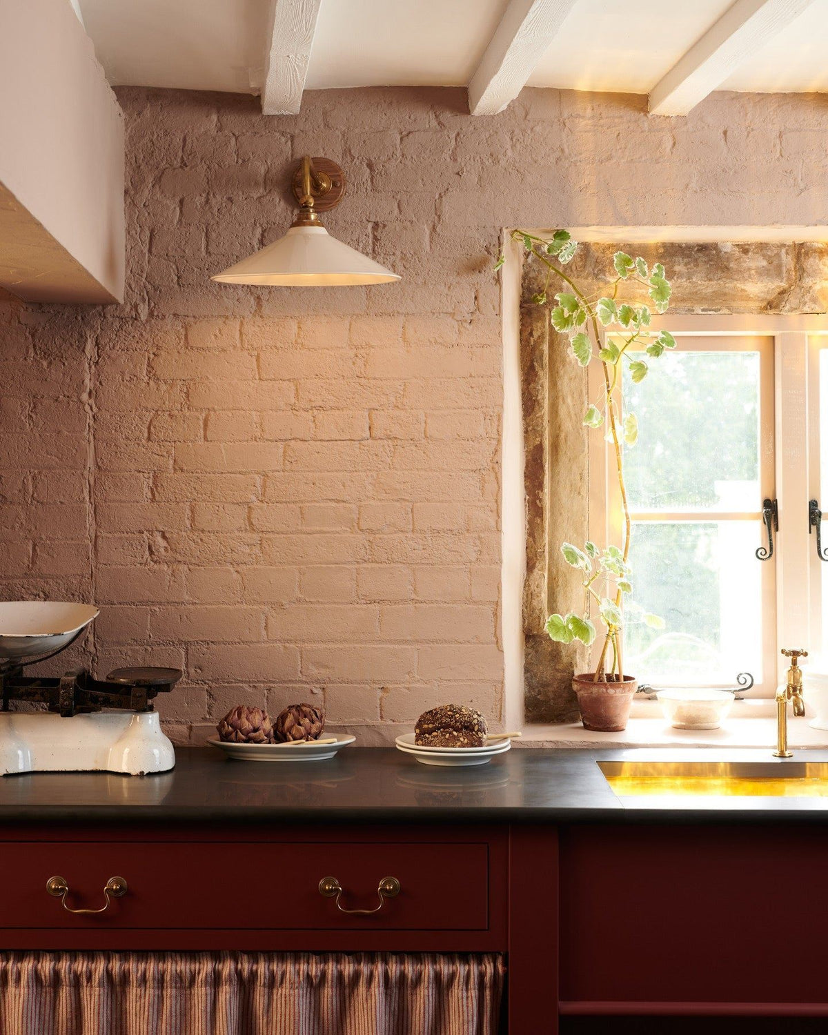 A straight-on view of a large Victorian-inspired wall light with a brass arm and a cream glazed ceramic shade fixed to a pink painted brick wall above a slate worktop and red painted cabinetry. TEST