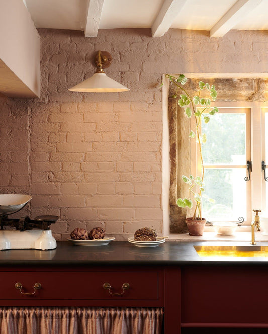 A straight-on view of a large Victorian-inspired wall light with a brass arm and a cream glazed ceramic shade fixed to a pink painted brick wall above a slate worktop and red painted cabinetry.