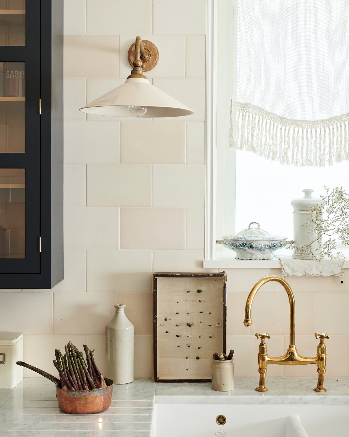 A straight-on view of a large Victorian-inspired wall light with a brass arm and a cream glazed ceramic shade fixed to a wall of cream subway tiles above a kitchen sink. TEST