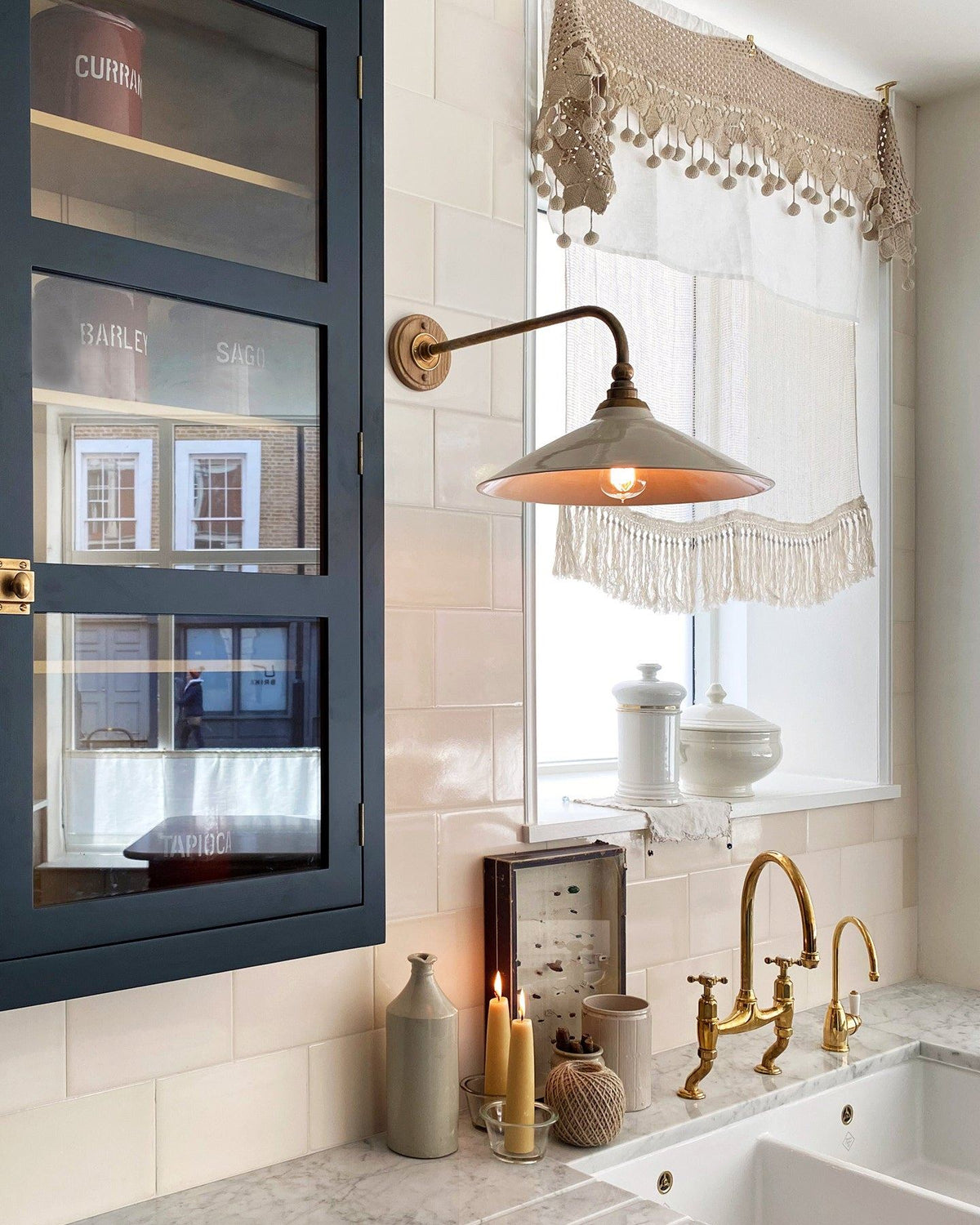 An oblique view of a large Victorian-inspired wall light with a brass arm and a cream glazed ceramic shade fixed to a wall of cream subway tiles above a kitchen sink. TEST