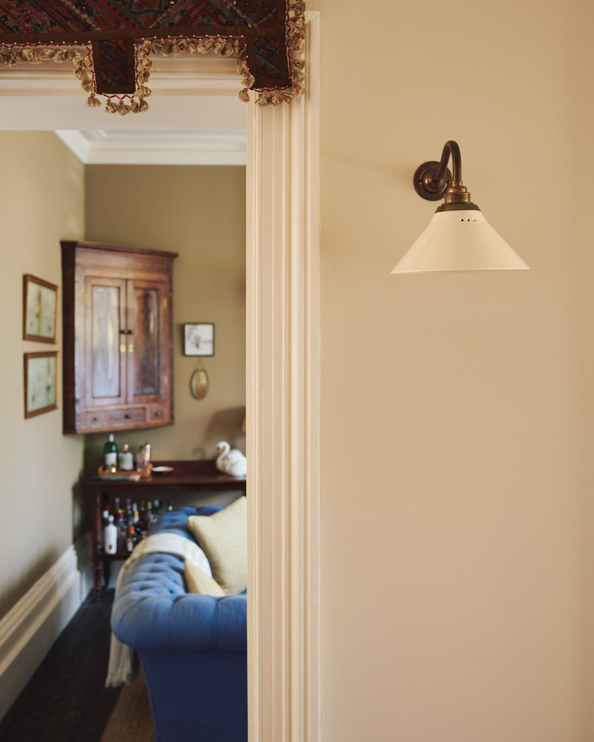 An oblique view of a small wall light with an aged brass swan neck arm and a small cream glazed ceramic shade, fixed to a cream wall. TEST