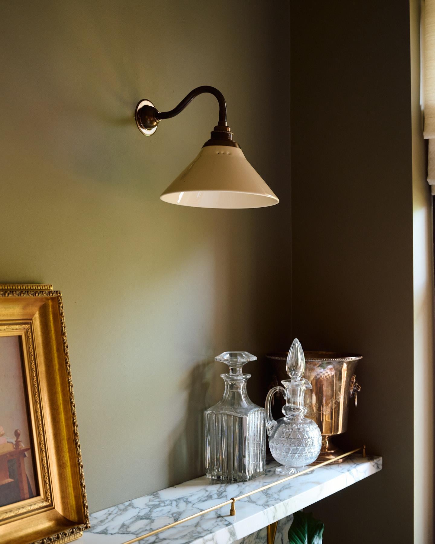 An oblique view of a small wall light with an aged brass swan neck arm and a small cream glazed ceramic shade, fixed to a pink brick wall beside a linen curtain hanging from an aged brass rail.