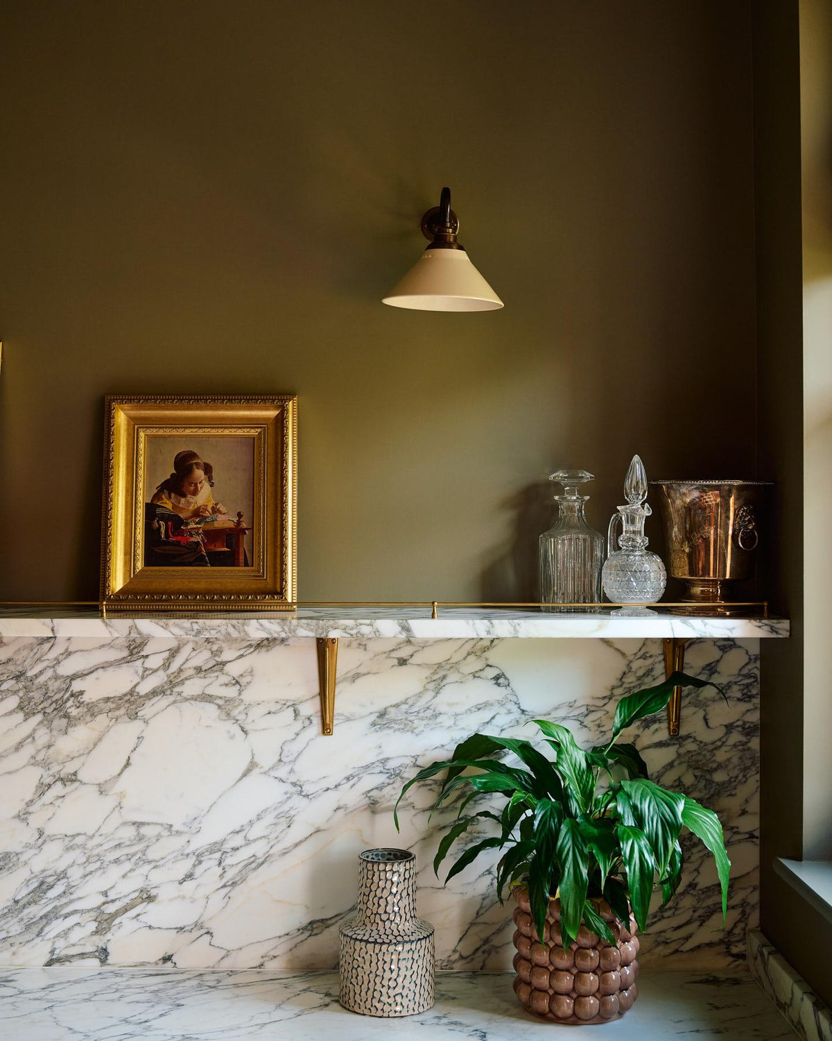 An oblique view of a small wall light with an aged brass swan neck arm and a small cream glazed ceramic shade, fixed to a dark green wall above an Arabescato marble shelf with glass bottles. TEST