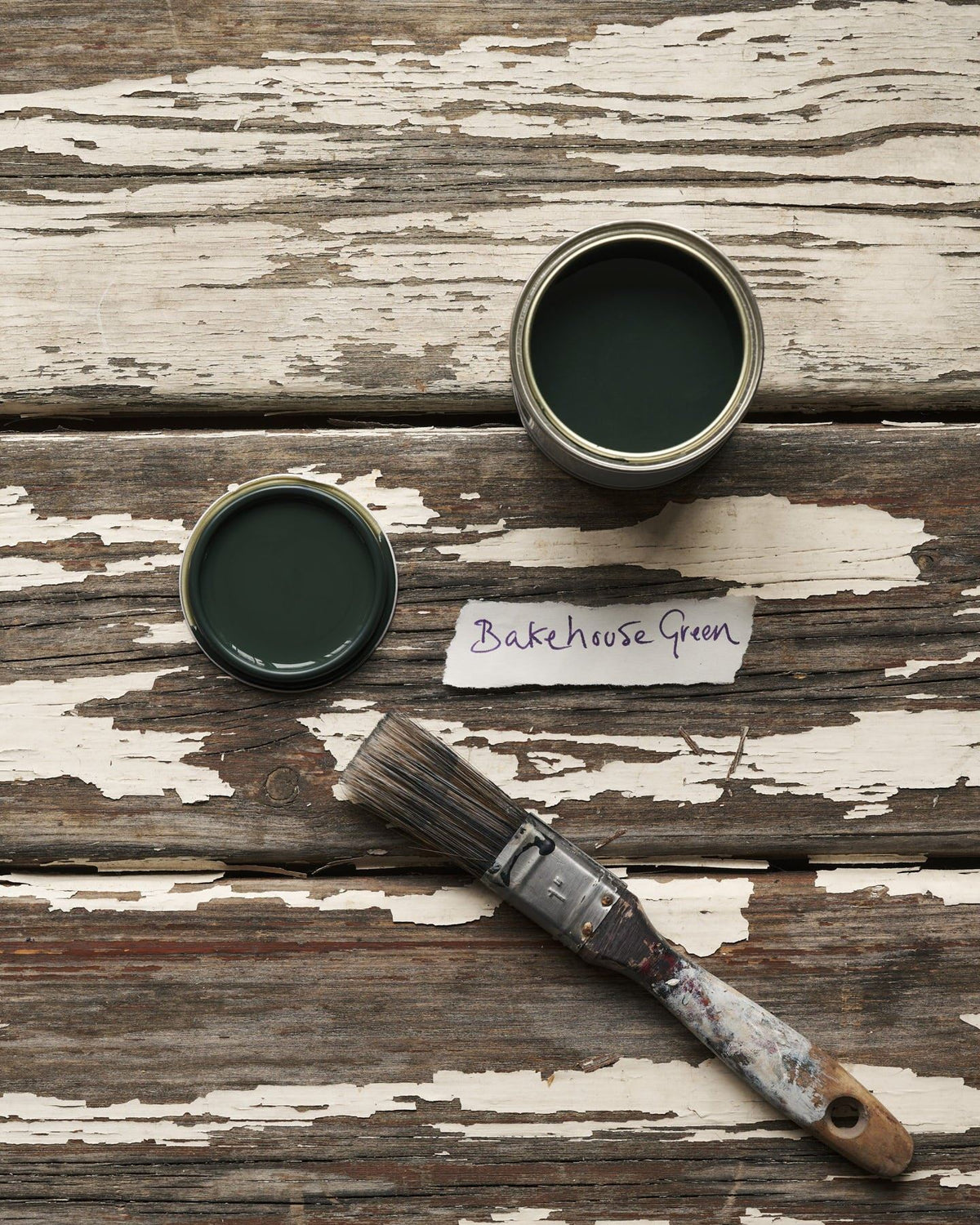 An overhead view of an open paint pot and a worn brush on a rustic tabletop. The dark green colour inside is labelled 'Bakehouse Green'. TEST