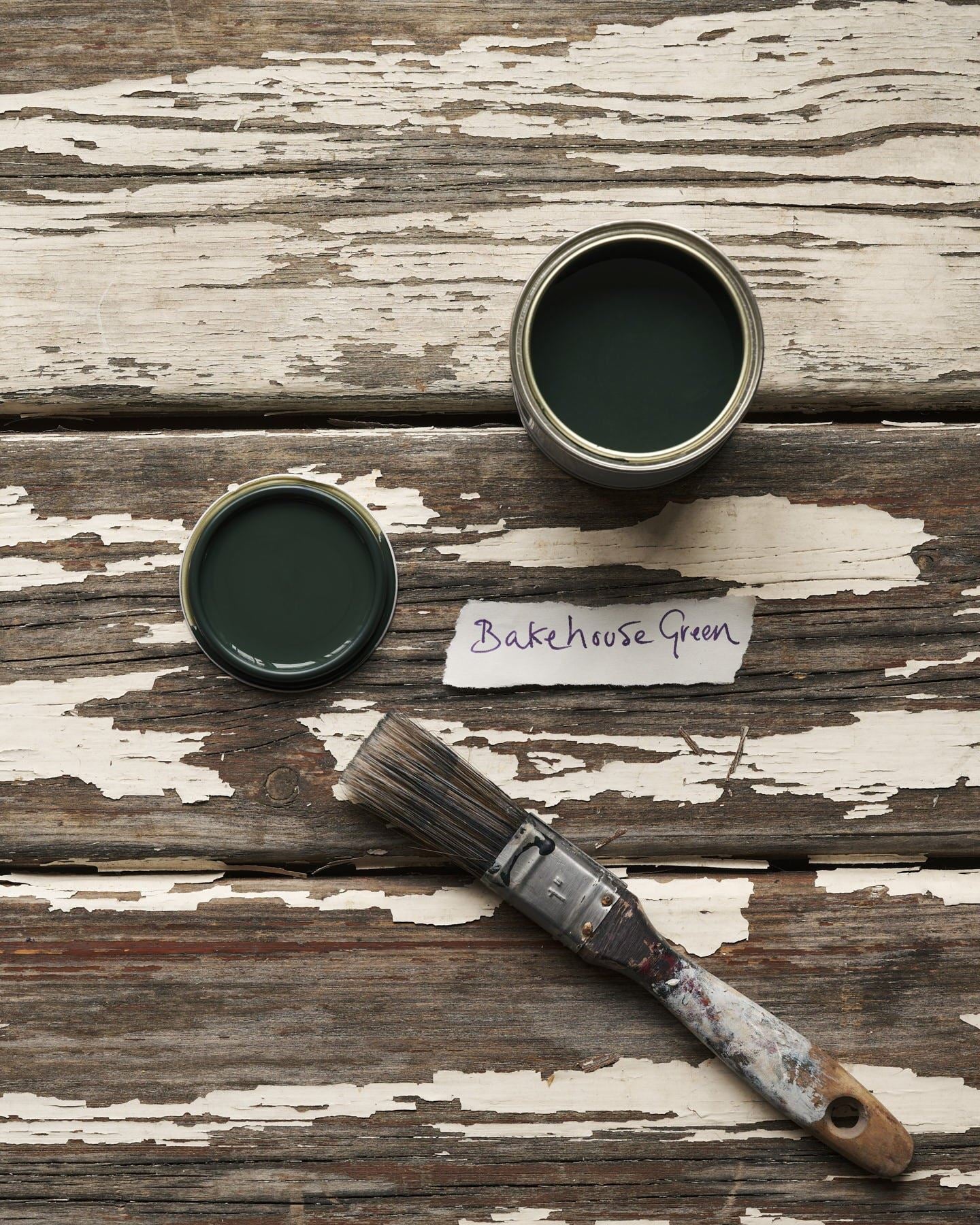 An overhead view of an open paint pot and a worn brush on a rustic tabletop. The dark green colour inside is labelled 'Bakehouse Green'.