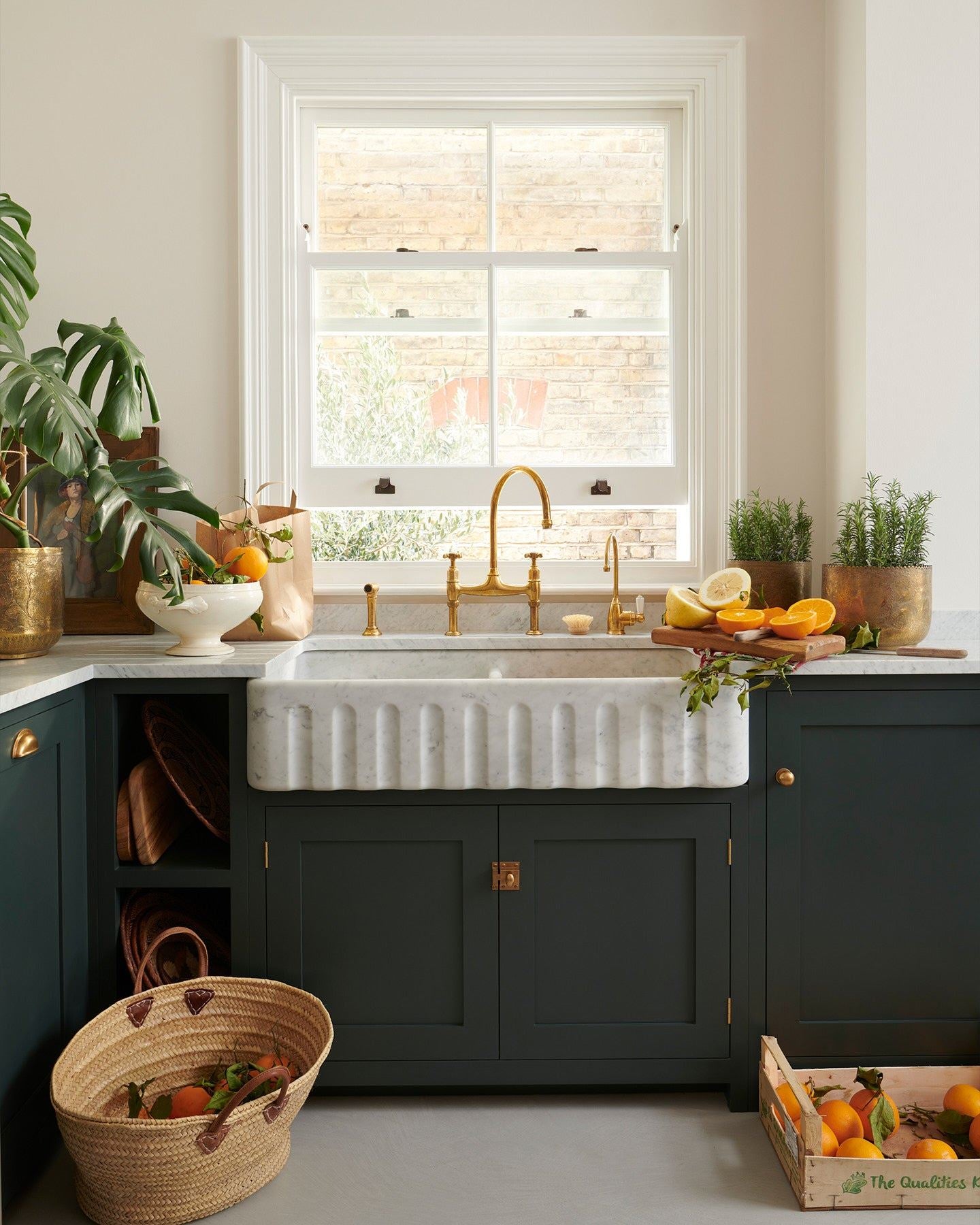 A dark green painted Shaker-style kitchen with light coloured walls, a cement floor, a wide Carrara marble sink with a fluted front, aged brass taps, a monstera plant and baskets of vibrant oranges.