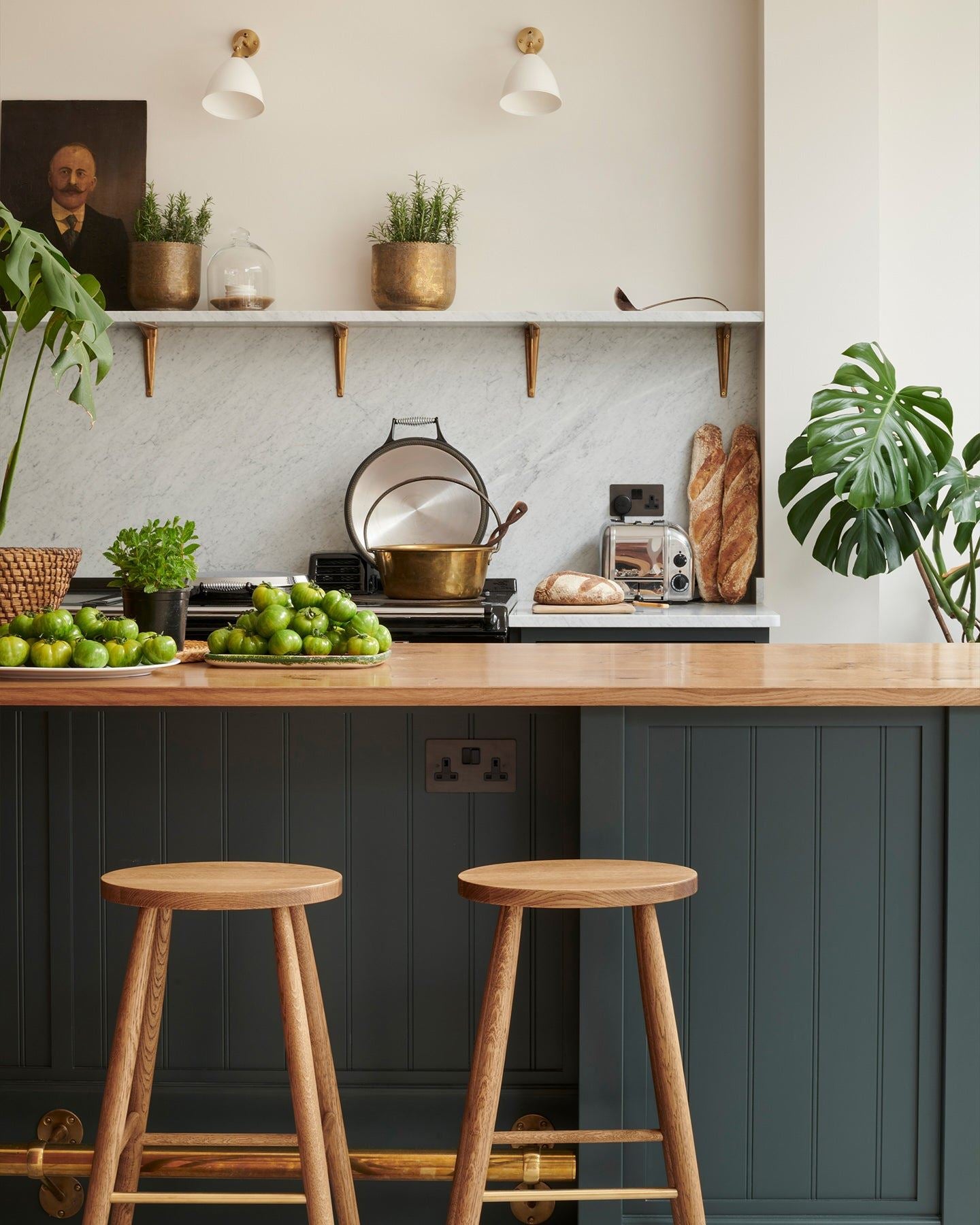 A dark green painted Shaker-style kitchen with a breakfast bar island, simple oak bar stools and a Carrara marble splashback.