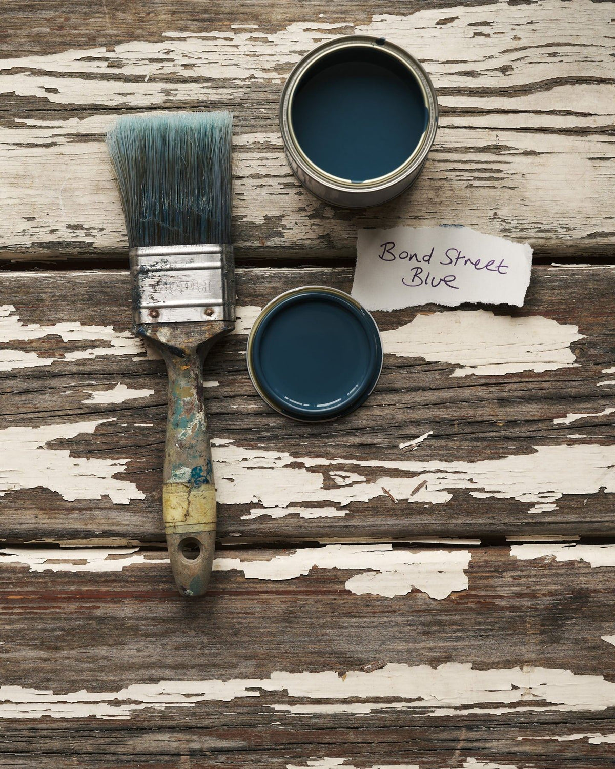 An overhead view of an open paint pot and a worn brush on a rustic tabletop. The royal blue colour inside is labelled 'Bond Street Blue'. TEST