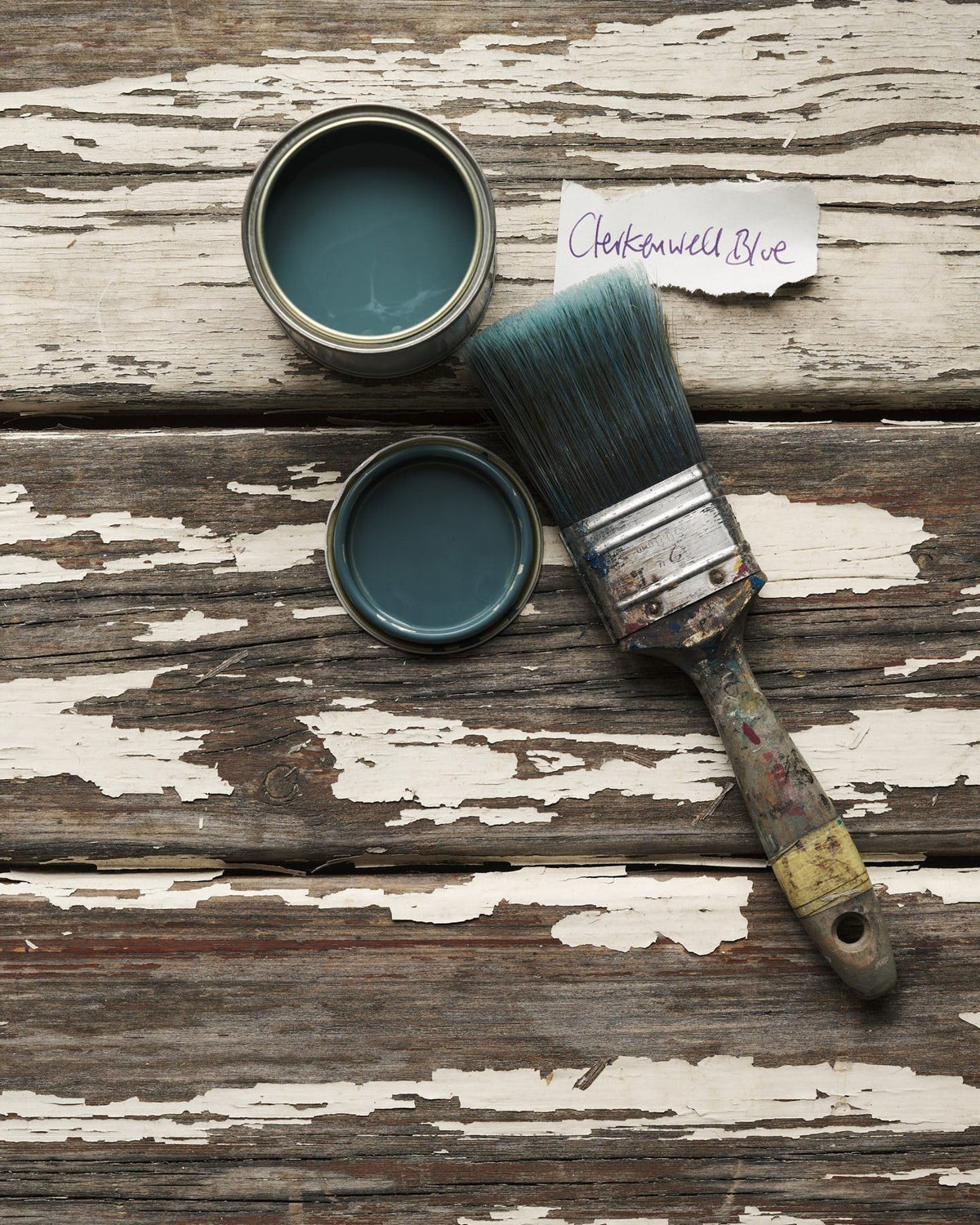 An overhead view of an open paint pot and a worn brush on a rustic tabletop. The green-tinged blue colour inside is labelled 'Clerkenwell Blue'. TEST