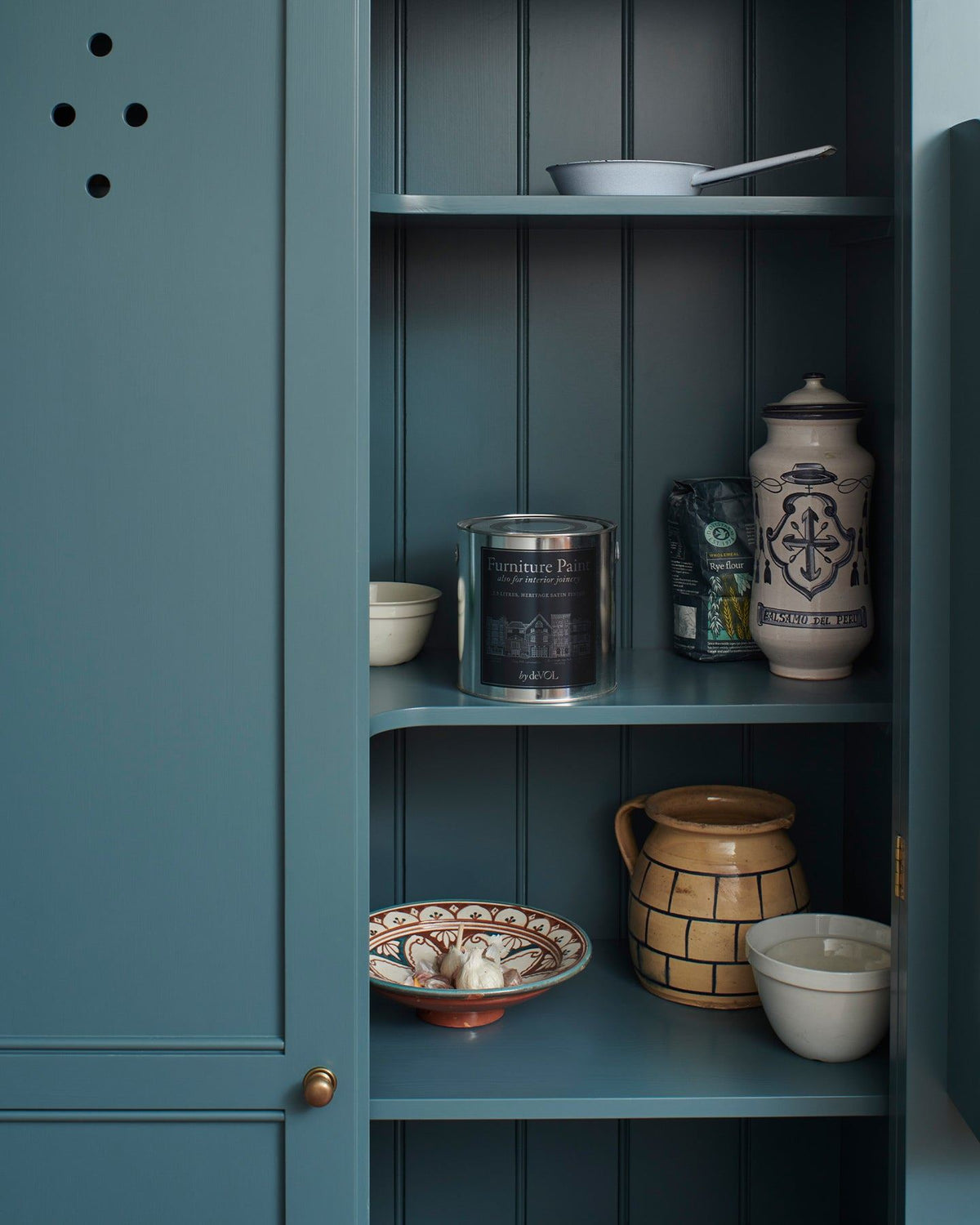 Looking into a Georgian-style pantry cupboard, painted inside and out in a green-tinged blue. Inside the cupboard there is a silver paint tin with a blue label and a collection of vintage crockery. TEST