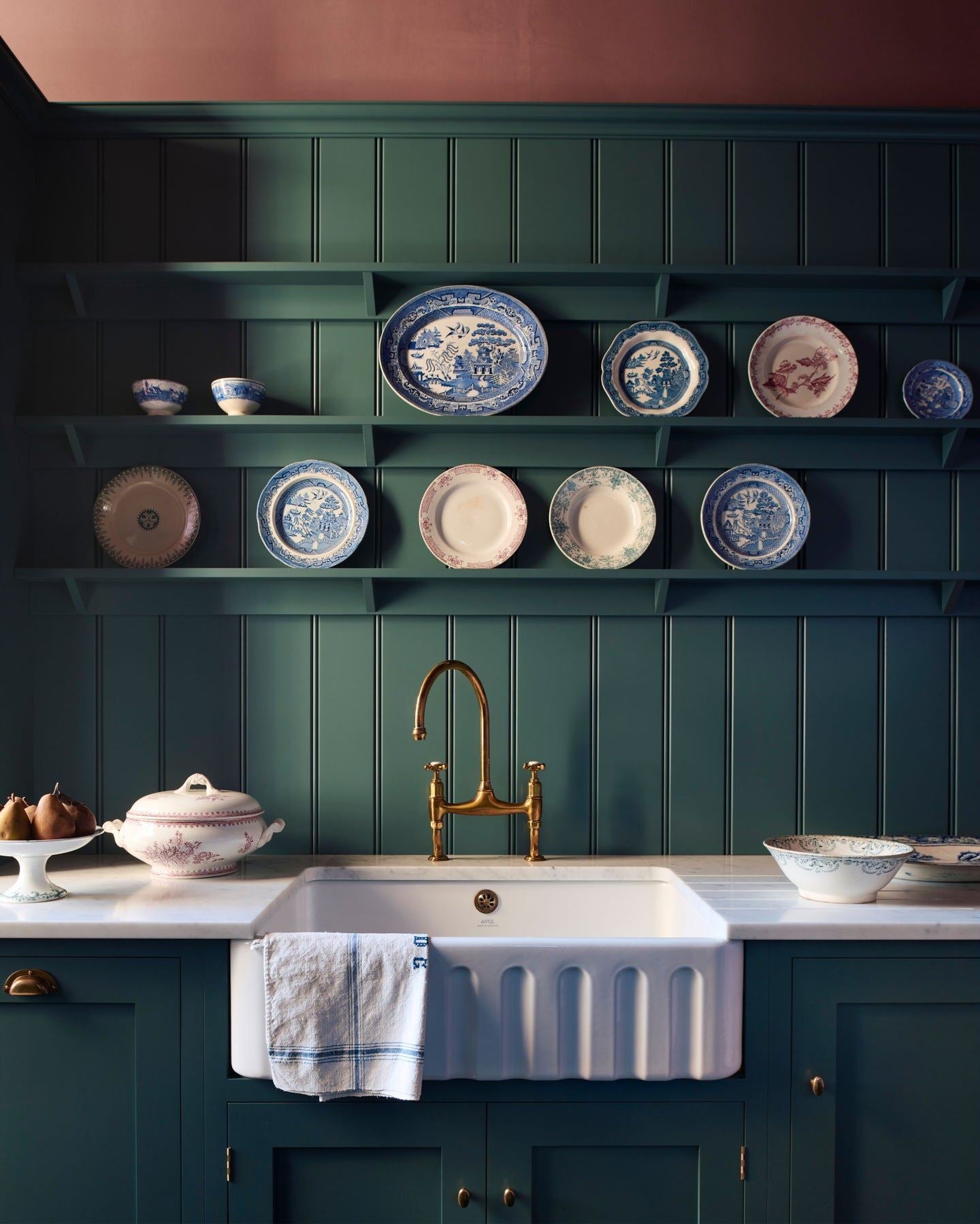 A straight-on view of a classically-styled kitchen sink with green-blue painted Shaker-style cupboards, tongue and groove wall panelling, shelving full of vintage crockery and wine red coloured walls.