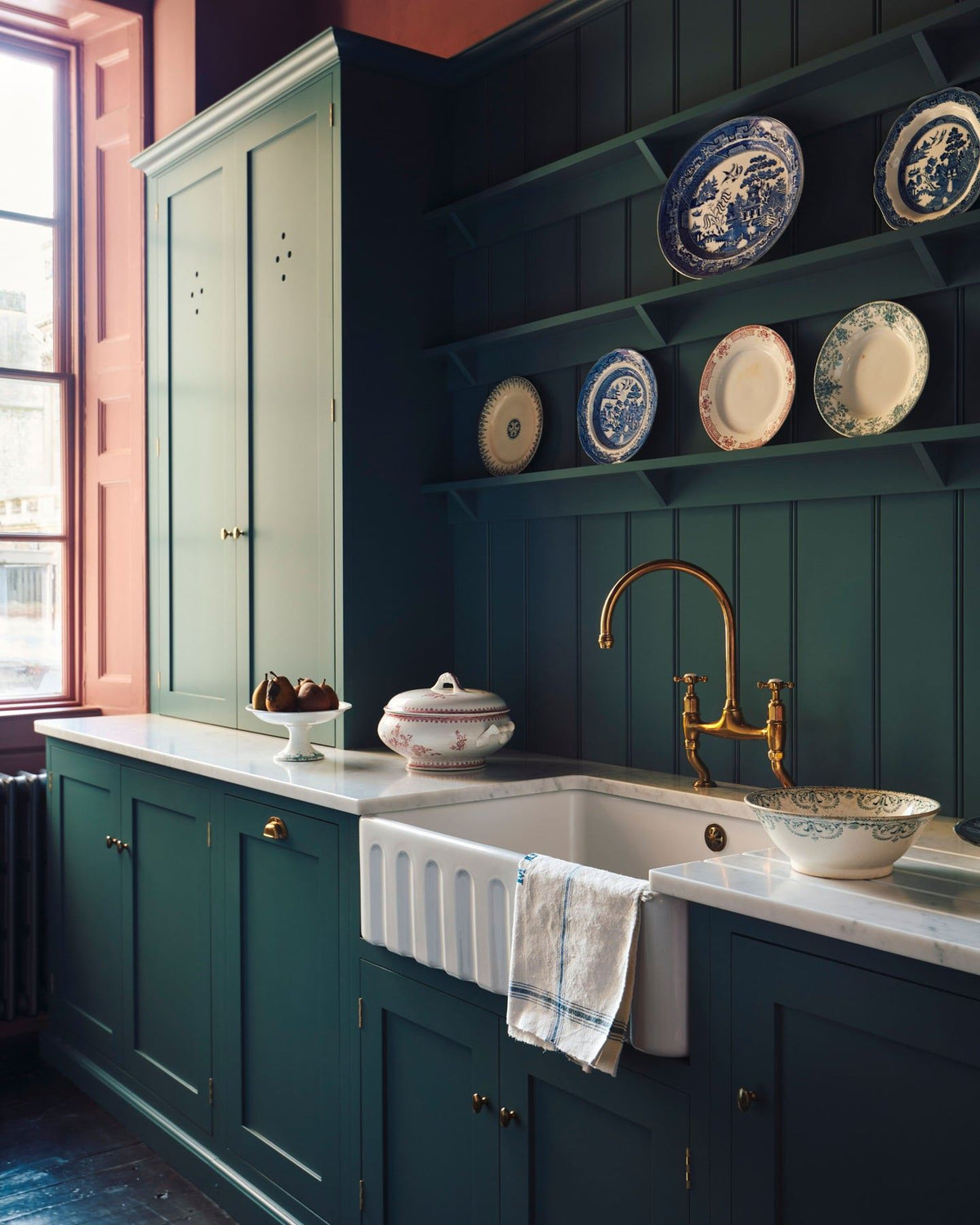 An oblique view of a classically-styled kitchen sink with green-blue painted Shaker-style cupboards, tongue and groove wall panelling, shelving full of vintage crockery and wine red coloured walls. TEST