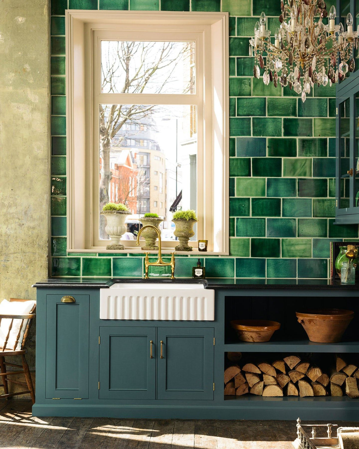 A straight-on view of a Georgian-style kitchen sink cabinet painted in a rich green-blue, with a black granite worktop, a wall of glossy emerald green tiles and an antique chandelier hanging high above. TEST