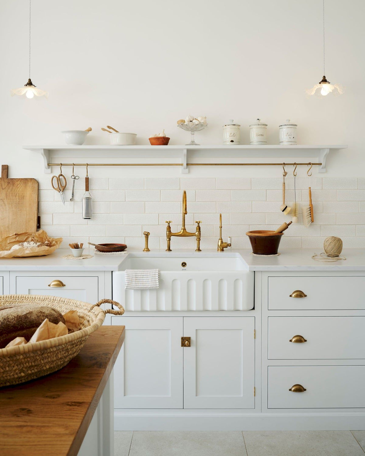 A straight-on view of a simple Shaker-style kitchen with off-white painted cupboards, a fluted Belfast sink, aged brass taps and white metro tiles. TEST