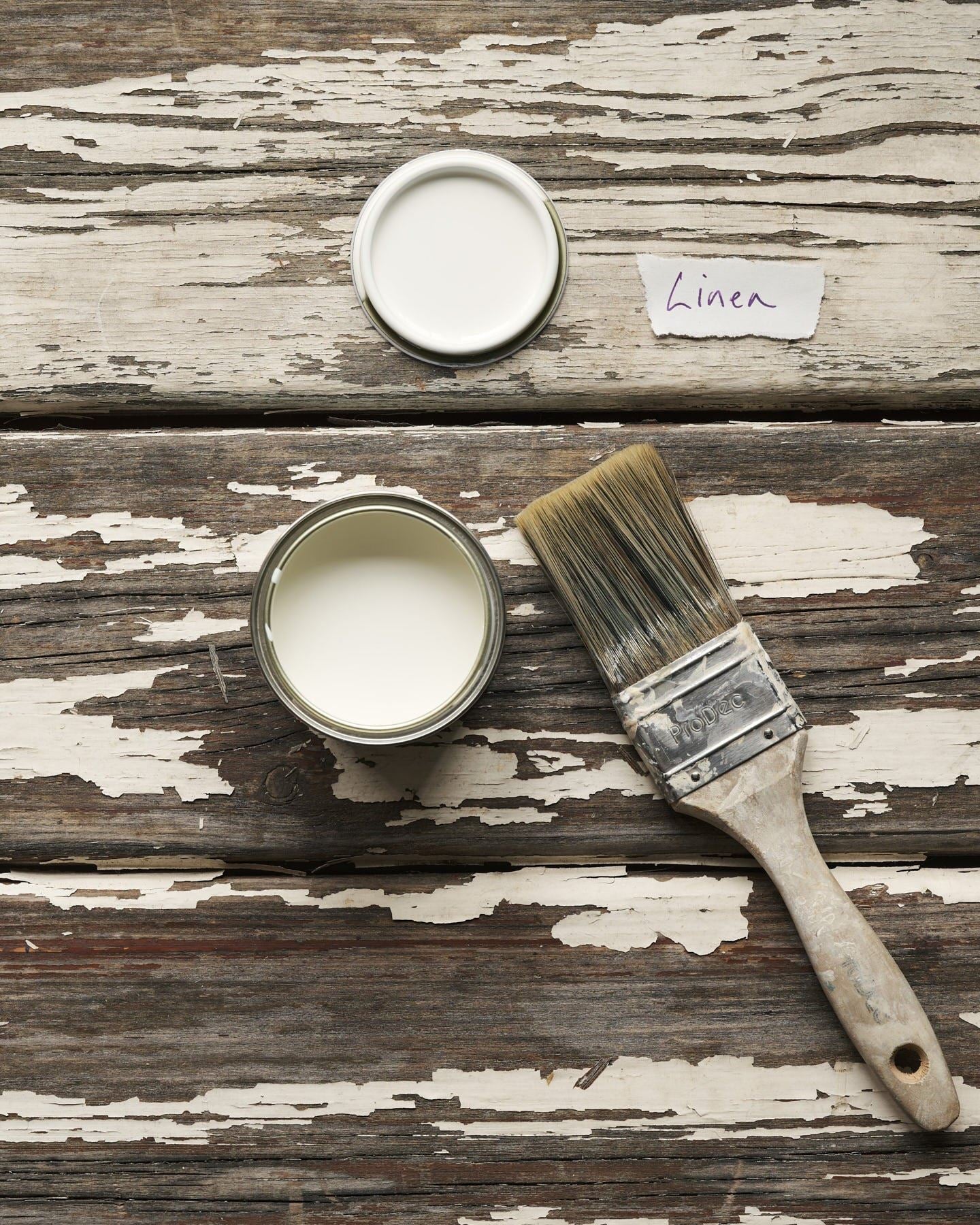 An overhead view of an open paint pot and a worn brush on a rustic tabletop. The light cream colour inside is labelled 'Linen'.