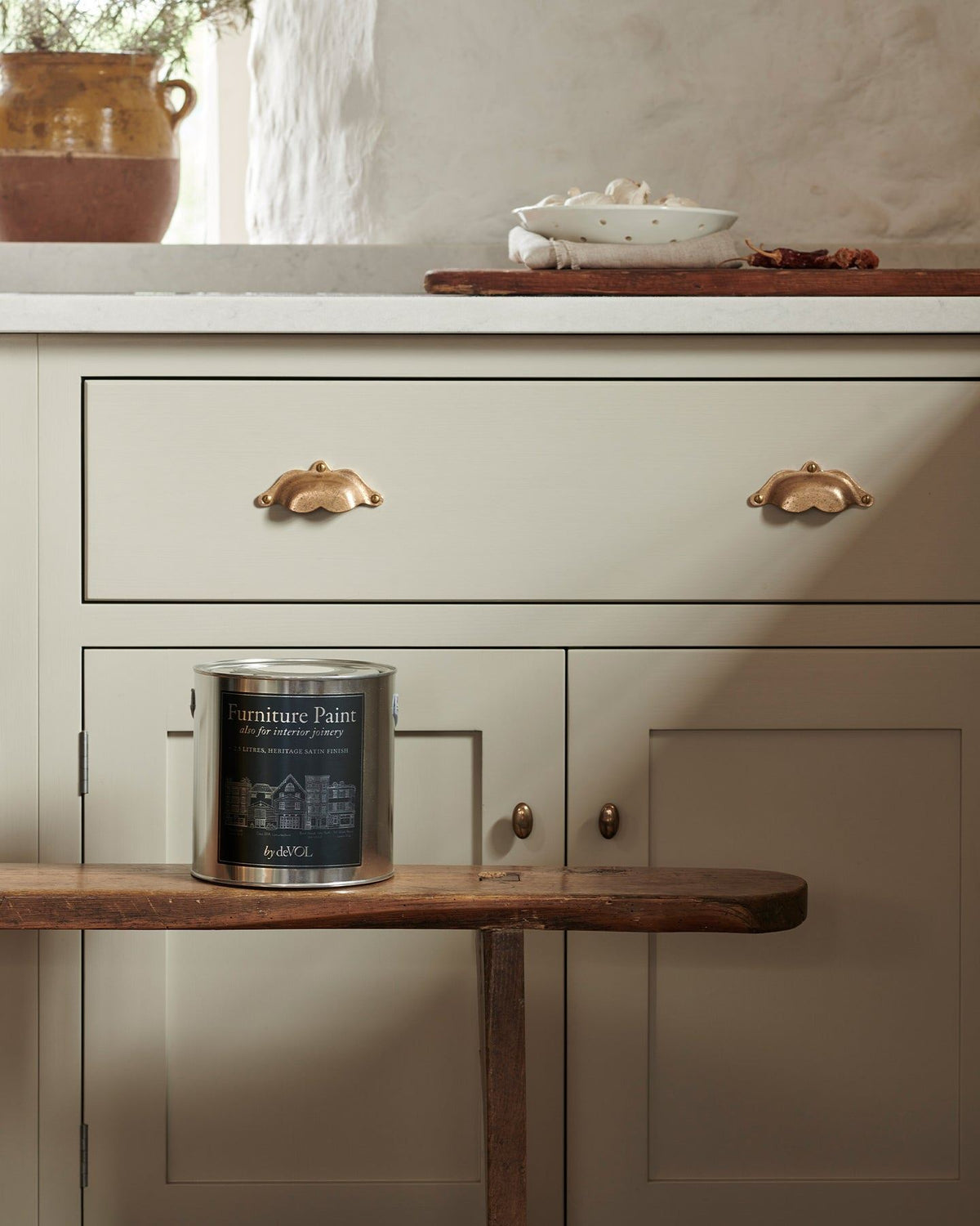 A silver paint tin with a blue label on an antique bench, in front of a mushroom coloured Shaker-style door/drawer base cabinet with ornate bronze pull handles. TEST
