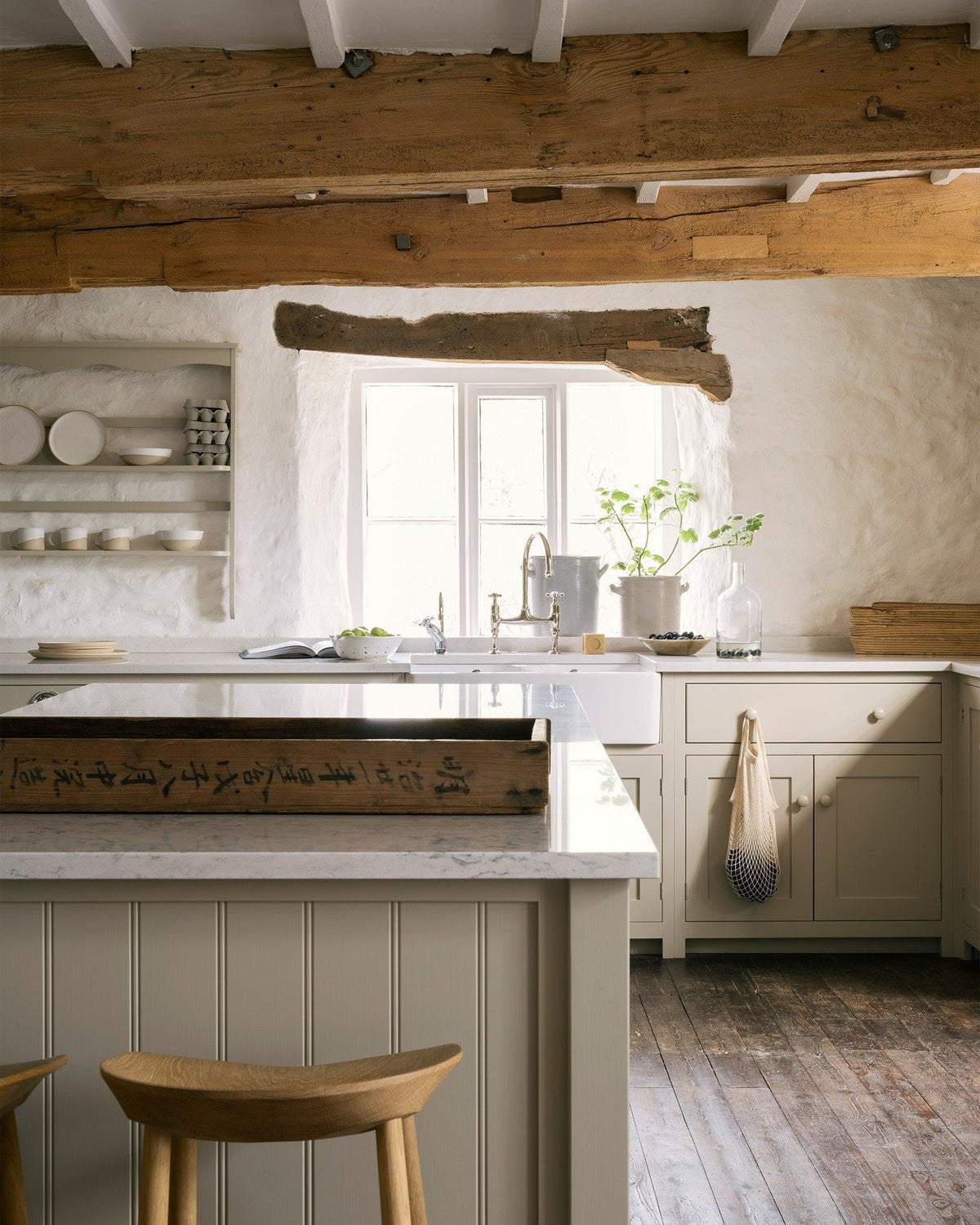 A large cottage kitchen with creamy mushroom coloured Shaker-style kitchen cupboards, dark worn floorboards and large wooden ceiling beams. TEST