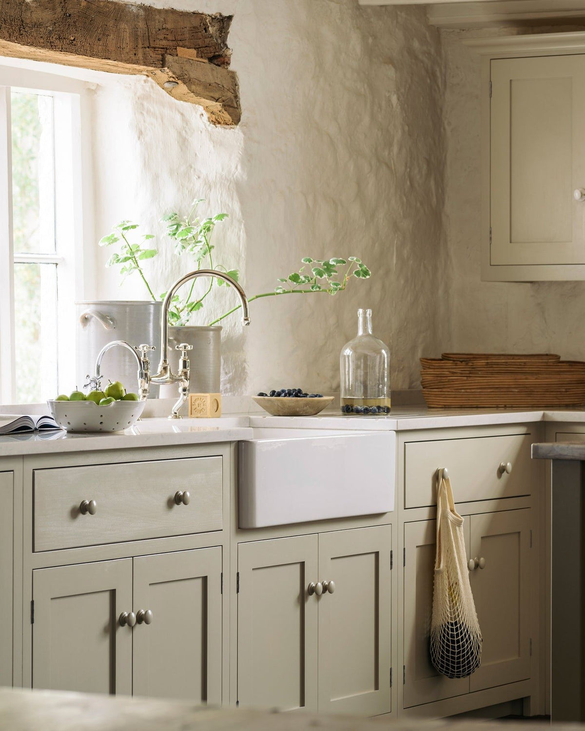 An oblique view of a Shaker-style kitchen sink cupboard, painted in a creamy mushroom colour and paired with chrome taps and a white ceramic Belfast sink. TEST