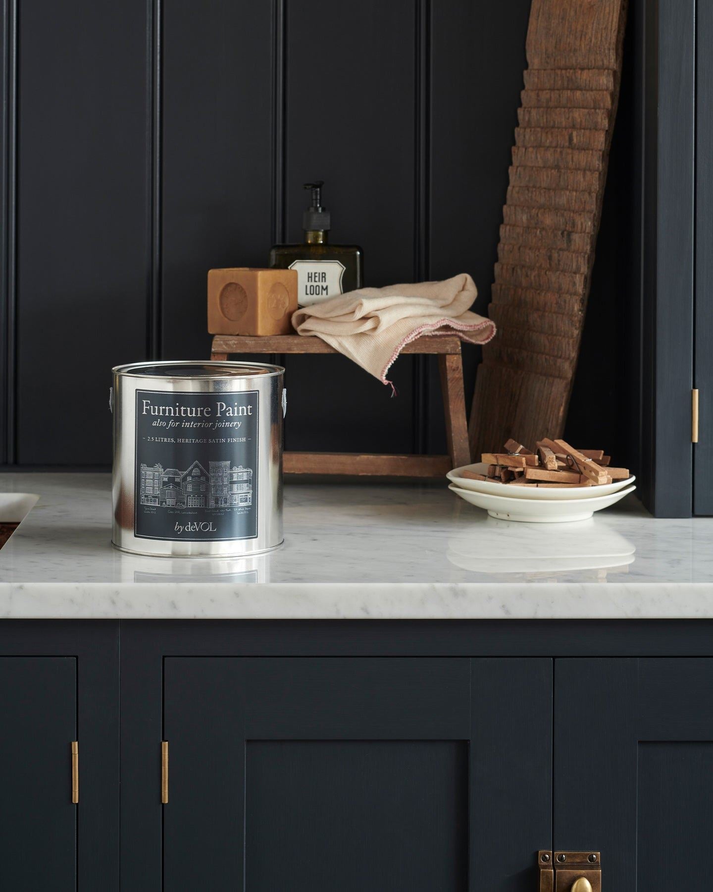 A close-up view of a silver paint tin with a blue label on a Carrara marble worktop, with dark blue painted cupboards and tongue and groove-style panelling behind.