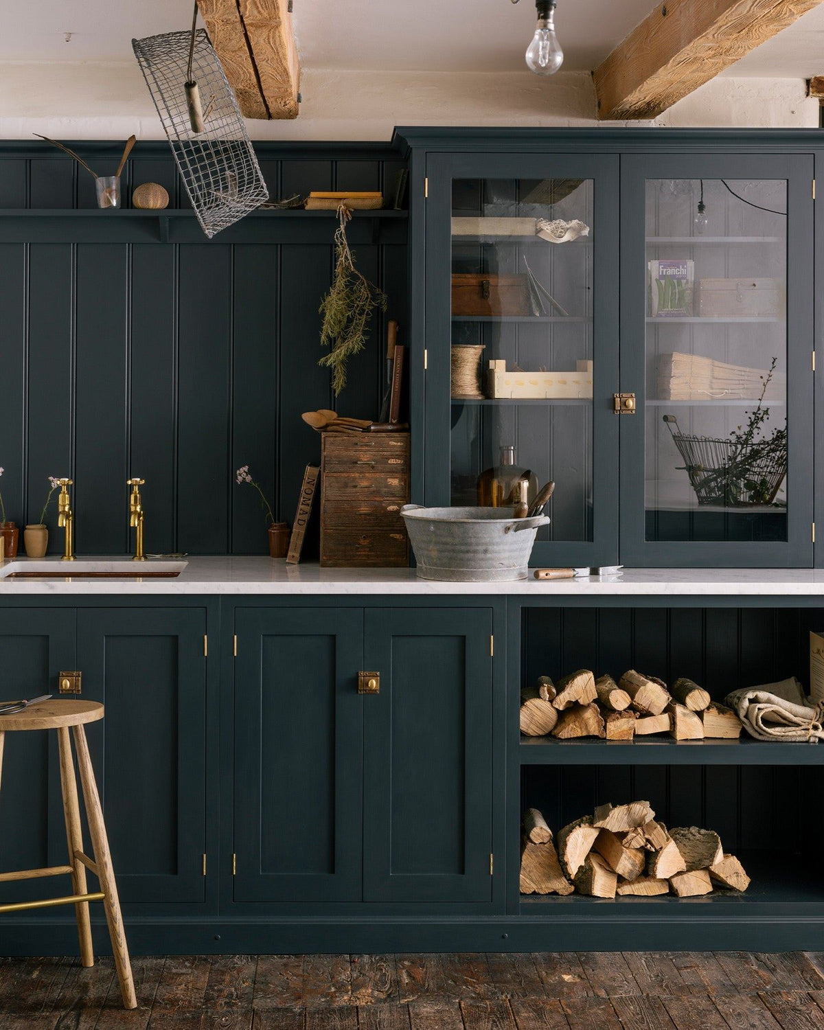 A straight-on view of a simple Shaker-style utility kitchen with dark blue painted cupboards, aged brass taps, tongue and groove-style panelling and a glazed countertop cupboard. The styling is rustic with logs piled in an open cupboard and a galvanised steel tub on the worktop. TEST