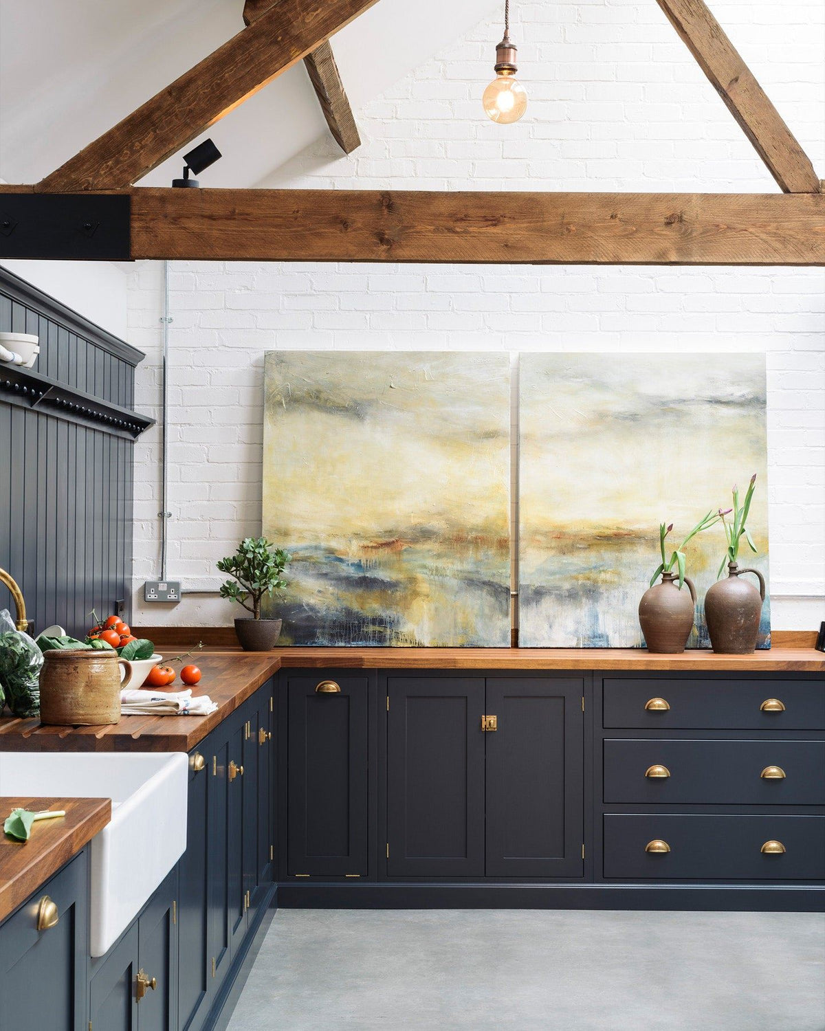 A large barn conversion with a simple Shaker-style kitchen, dark blue painted cupboards, brass hardware, rich hardwood worktops, white brick walls, a cement floor and old worn ceiling beams. TEST