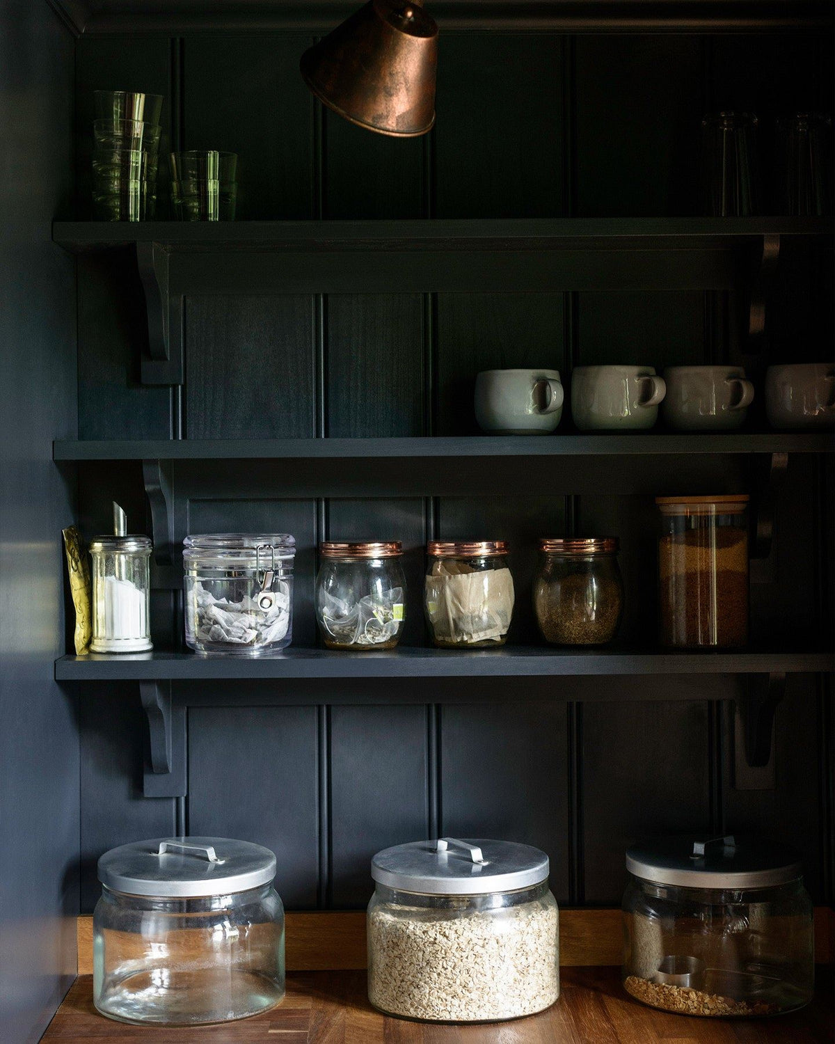 A dark blue painted tongue and groove panelled wall with Shaker-style shelving holding several jars of grains and cereals. TEST