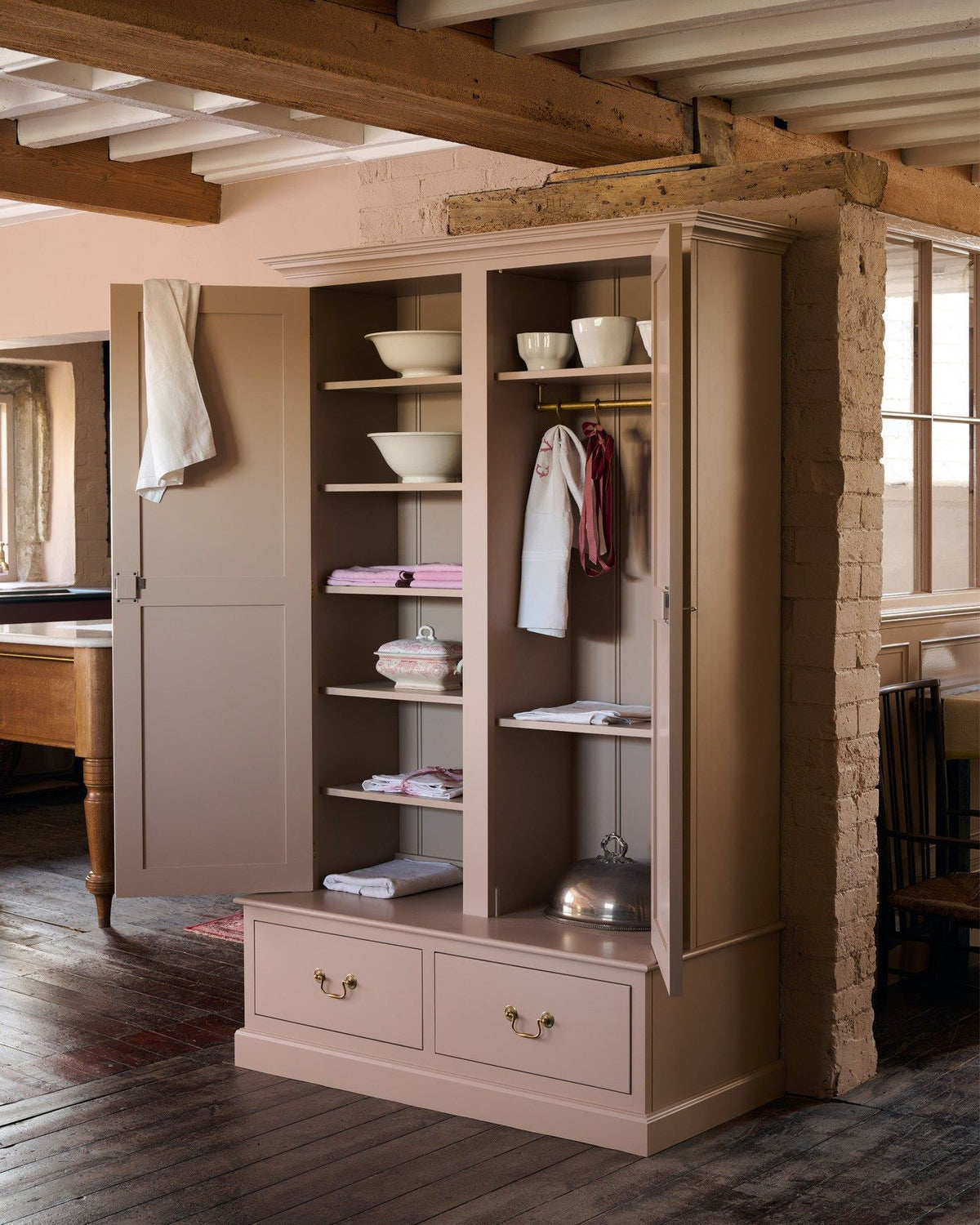 An oblique view of a Victorian-style pantry cupboard, painted inside and out in a soft pink colour. Both doors are open, revealing fixed shelving on the left and a tall hanging space on the right. A pedestal base contains two box drawers with Georgian-style brass pull handles. TEST