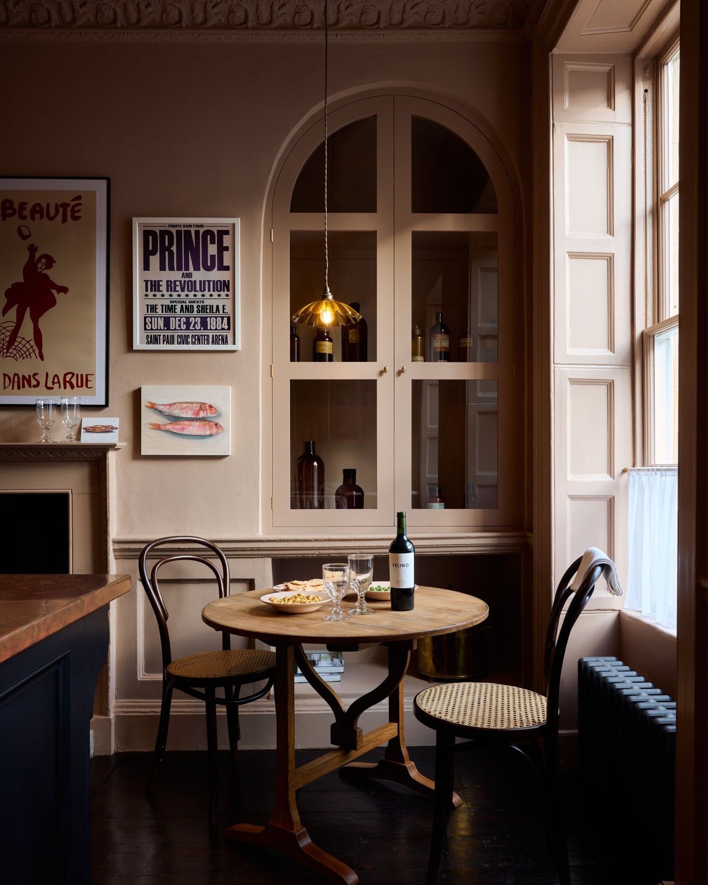 A straight-on view of a moody Georgian-style cafÈ-style corner with a small round pine dining table, pink walls and soft pink painted fitted glazed cupboard doors.